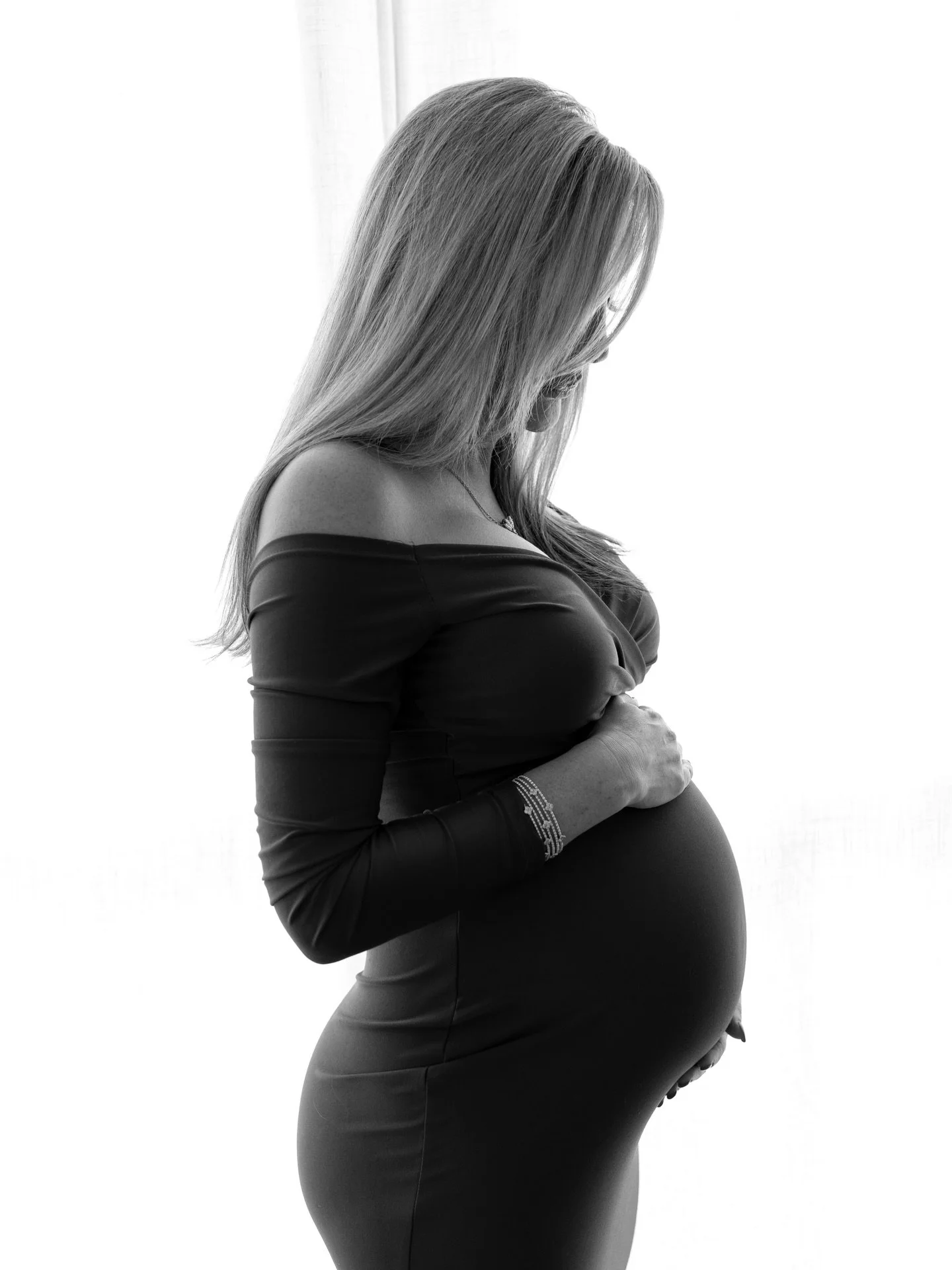 Black and white photo of a pregnant woman in a form-fitting dress, gently holding her belly and looking down.