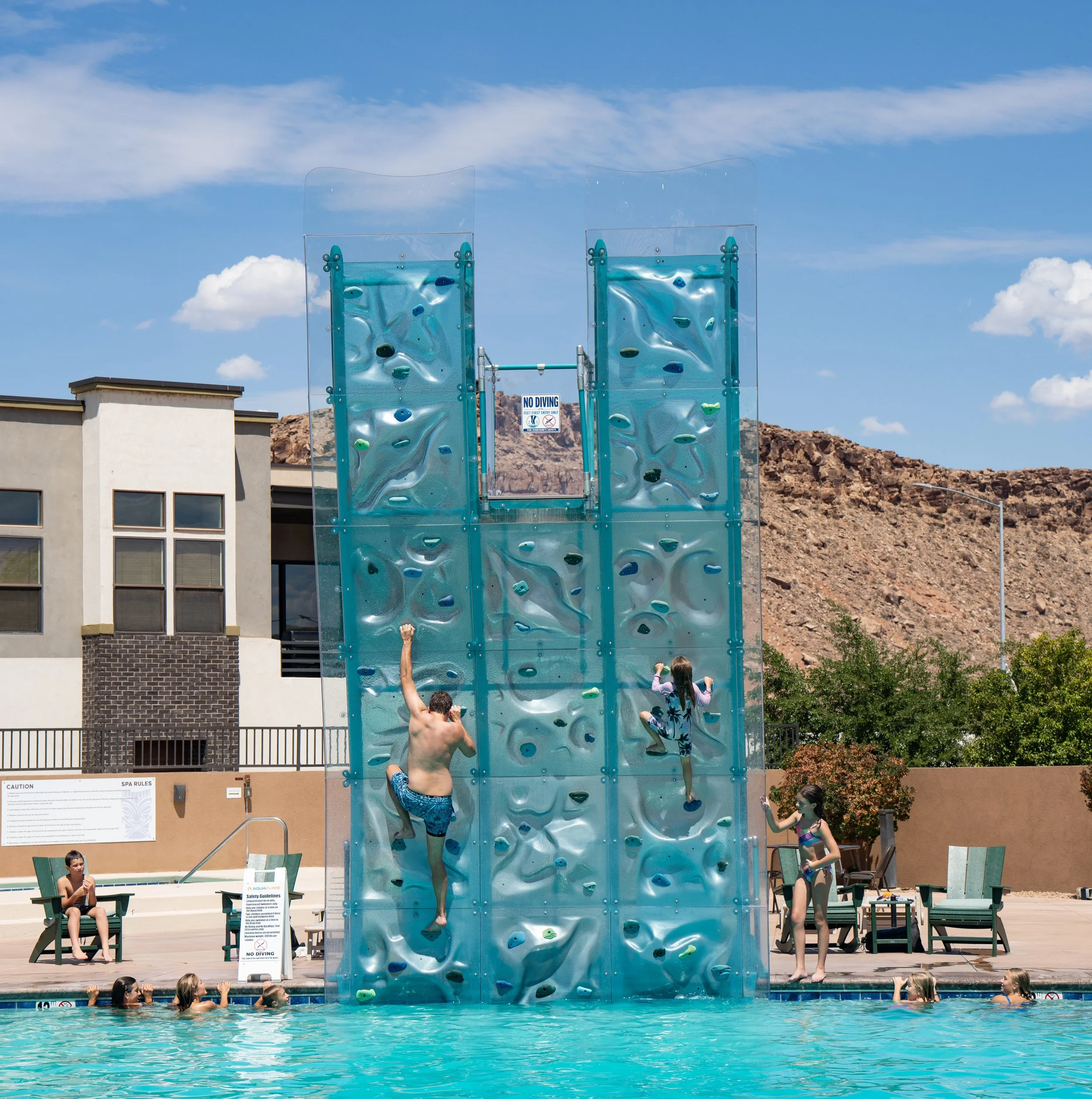 Children and adults at an outdoor swimming pool with a climbing wall, some people are swimming in the pool, others waiting or climbing on the wall, under a blue sky with clouds.