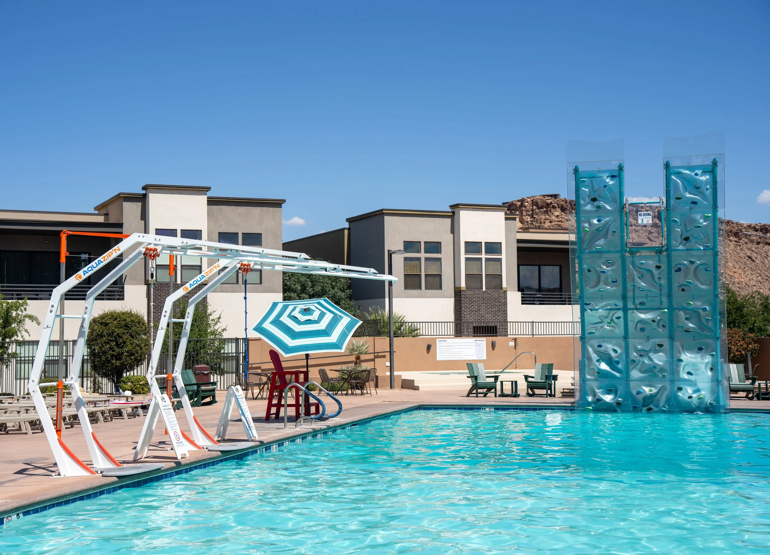Public swimming pool area with a water slide, climbing wall, shaded seating, and a modern residential building in the background under a clear blue sky.