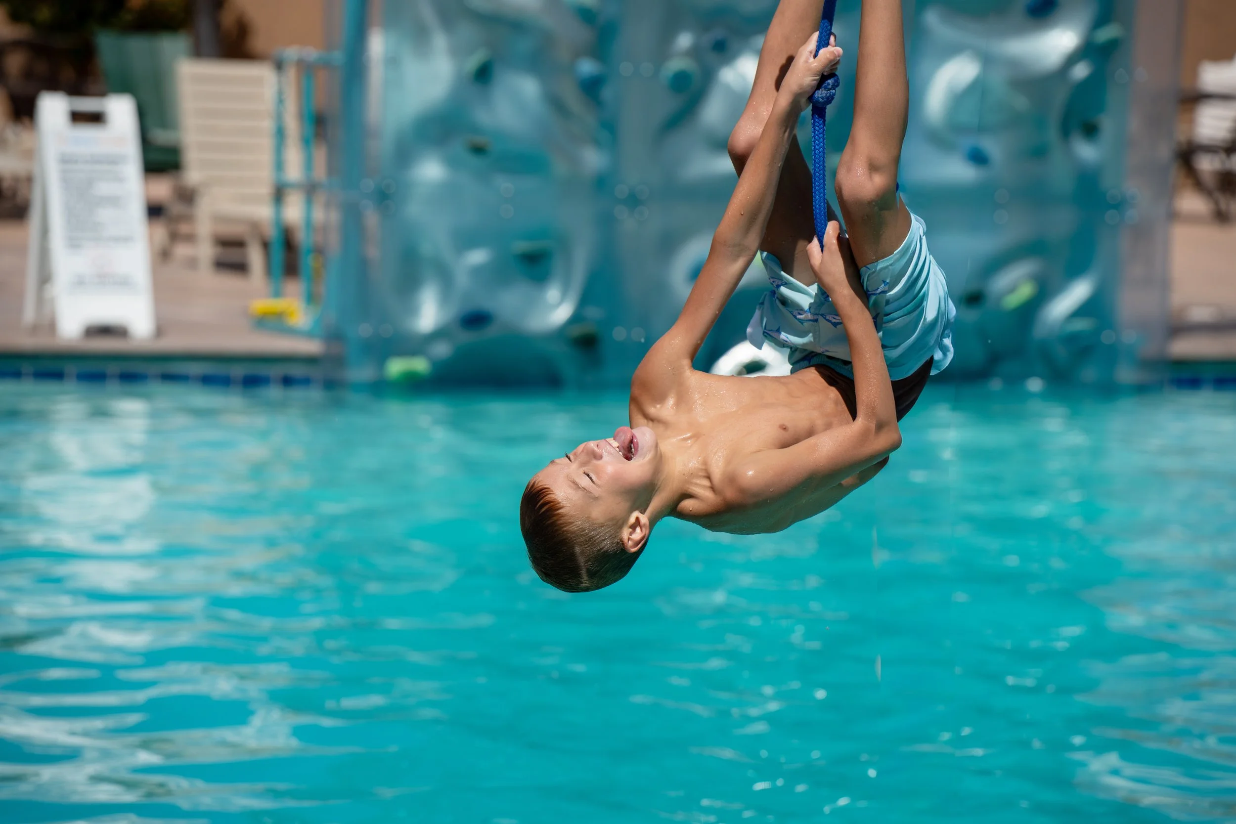 A boy falling backwards in a swimming pool on a rope zip and swing, with a  pool rock climbing wall and pool sign in the background.
