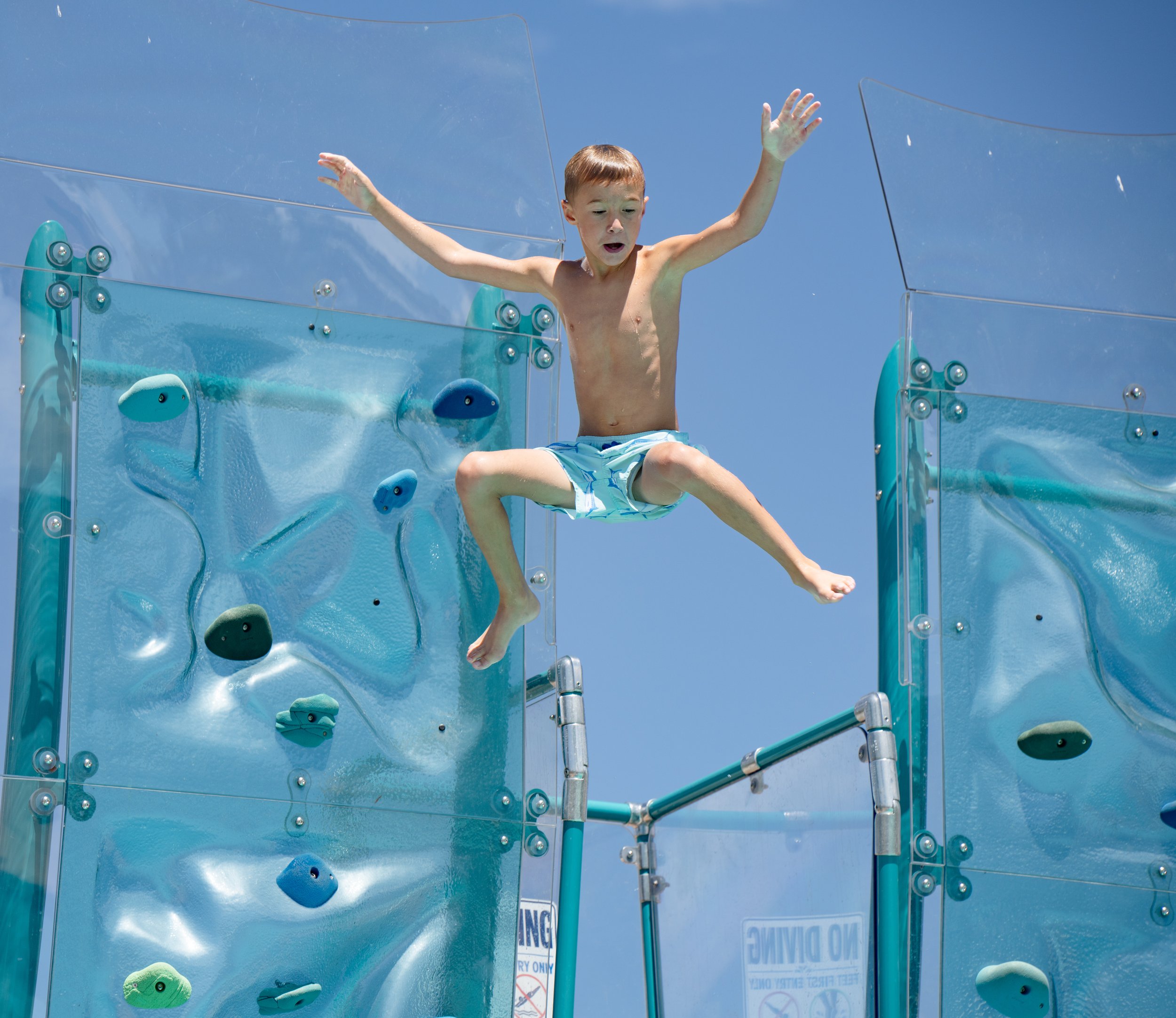 Young boy jumping form a platform on a pool climbing wall with holds, against blue sky, surprised expression.