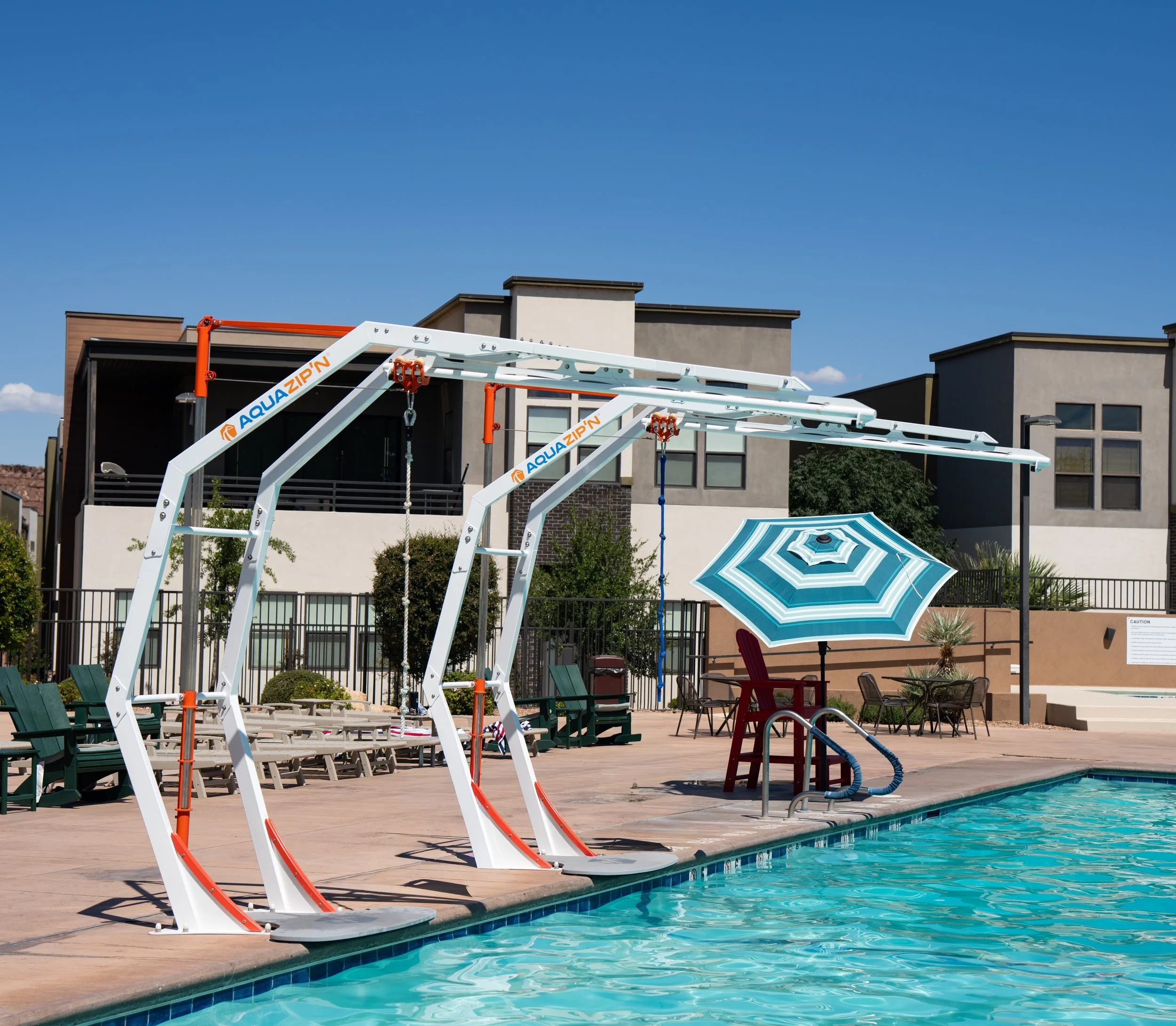 Swimming pool with poolside lounge chairs, umbrellas, and aquatic zip line equipment in a residential complex under a clear blue sky.