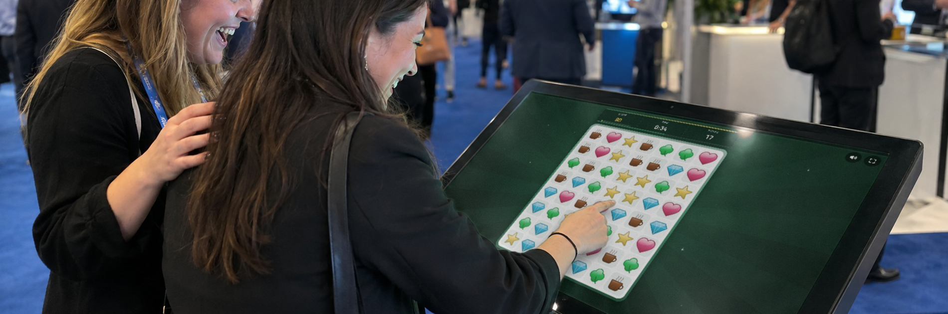 Branded puzzle game hire UK: two women laughing as they play Combo Craze, a match-3 game, on a touchscreen kiosk at a trade show