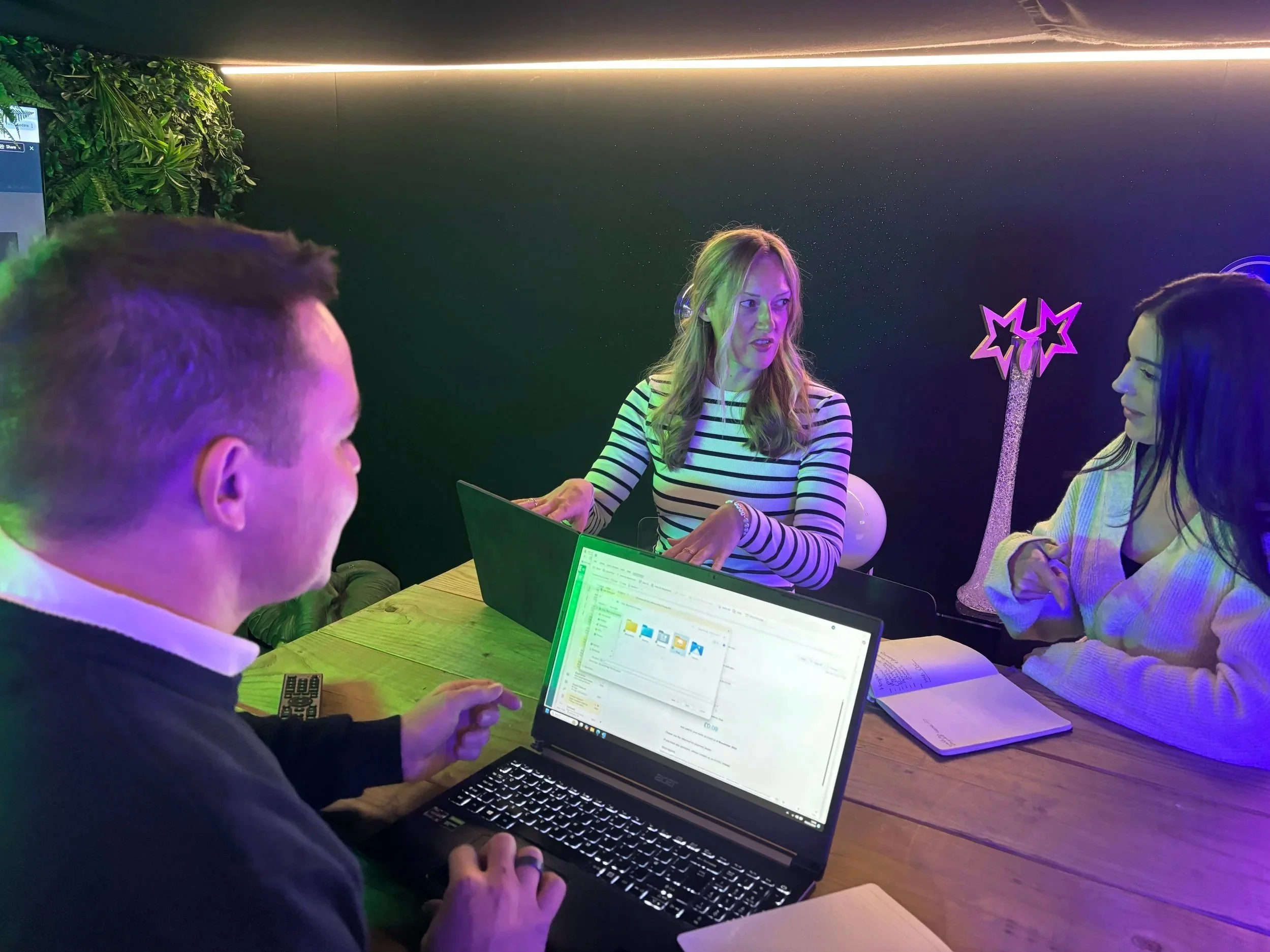 Three people in a client consultation at the Social Jungle Experiential Playground, reviewing a campaign presentation on a laptop in a green-lit showroom setting