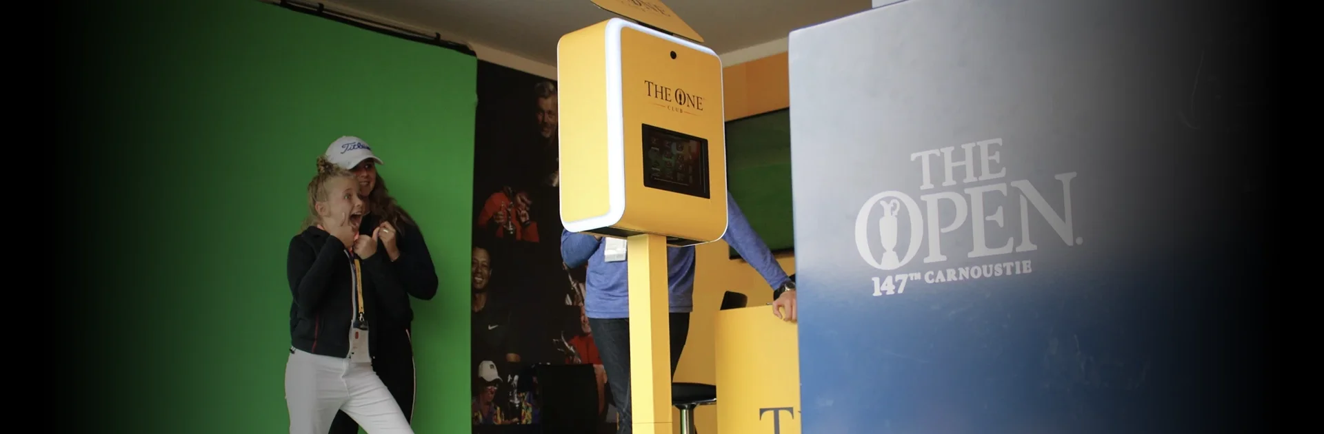 A young girl reacts with excitement as she sees her green screen photo result on a branded photo booth kiosk at The Open Championship 147th at Carnoustie, with a green screen backdrop visible behind her.