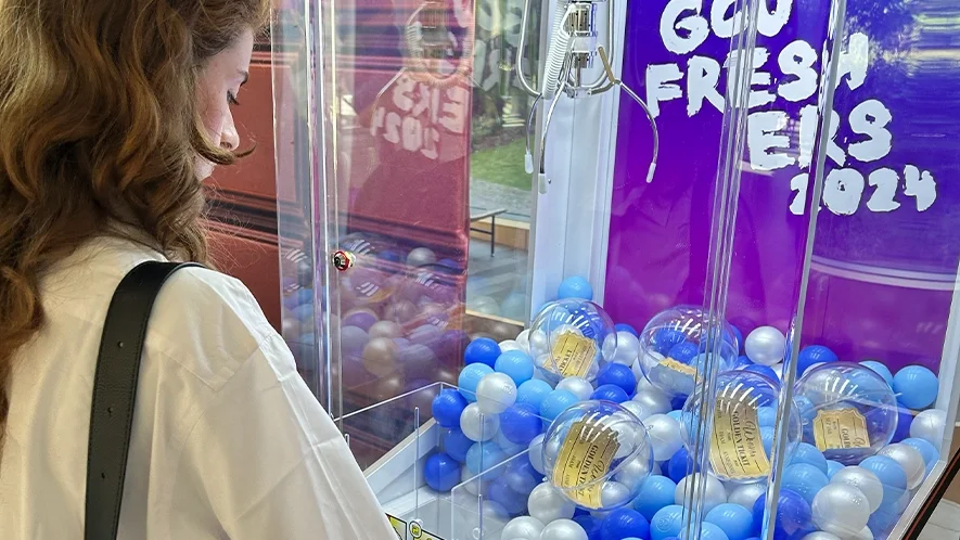 Student playing branded claw machine hire UK activation at Glasgow Caledonian University Freshers' Week 2024, cabinet filled with blue and pearl balls and Golden Ticket prize capsules