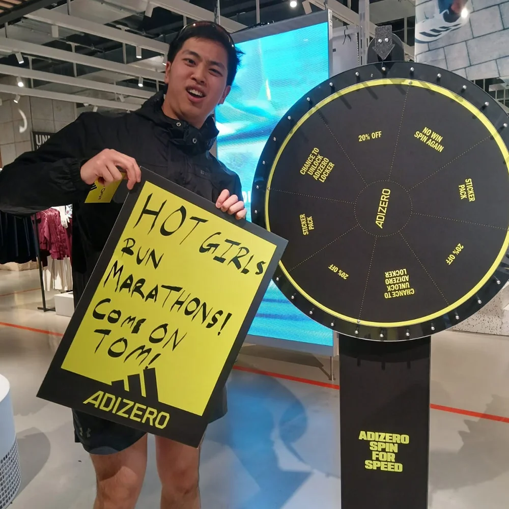 A shopper posing with a handmade ADIZERO sign beside the spin-to-win wheel at the Adidas Lockbox activation.