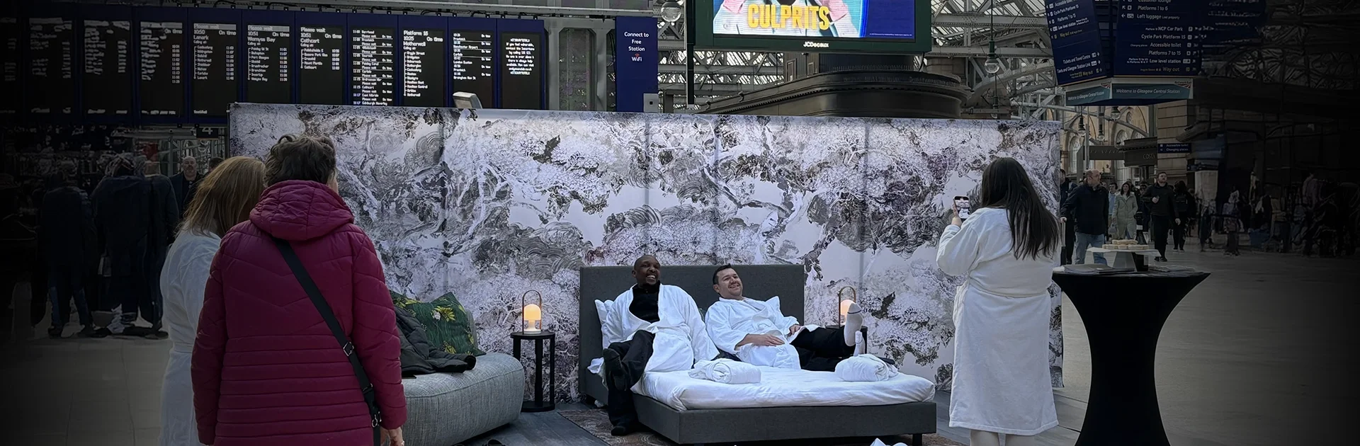Two men in white hotel robes relax on a Radisson Blu hotel bed, set up in the middle of Glasgow Central Station concourse, with a large marble-effect printed backdrop behind them. A woman in a white robe photographs them while commuters look on.