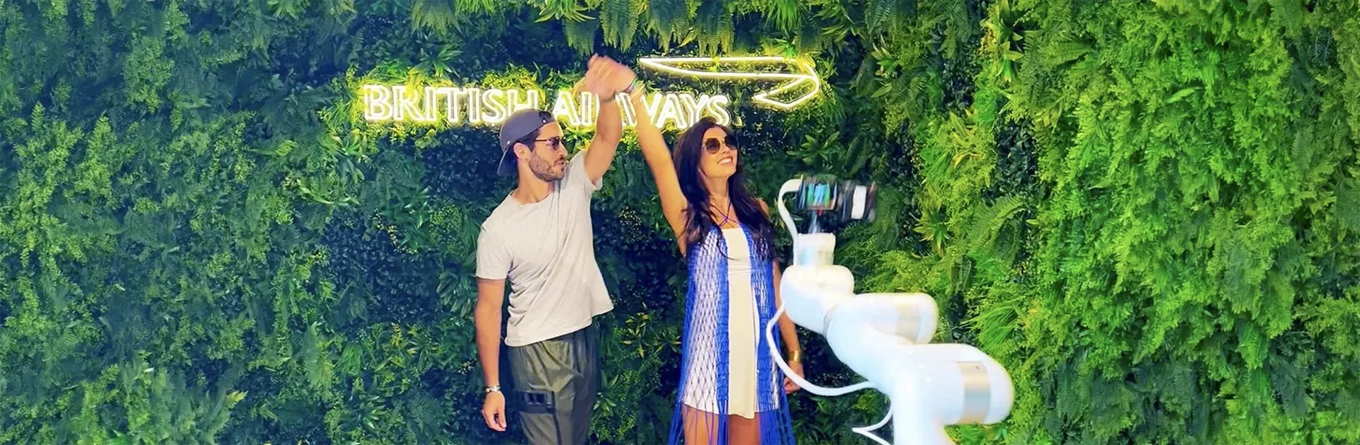 A man and woman pose and dance in front of a lush green living wall featuring a British Airways neon sign, while a white robotic camera arm films them at BST Hyde Park.