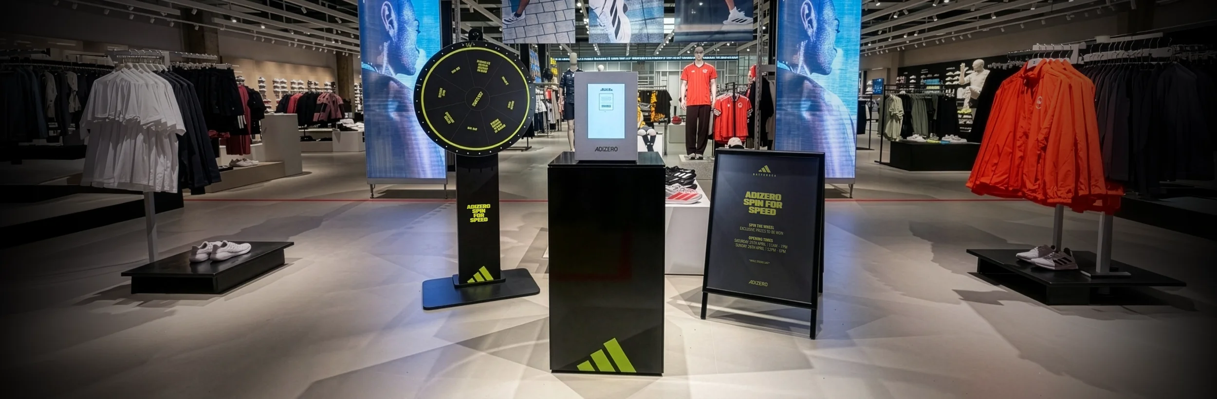 Three Adidas ADIZERO Spin for Speed branded activation units — a spin wheel, prize-reveal lockbox containing a pair of ADIZERO trainers to be won, and an A-board — displayed in an Adidas retail store.