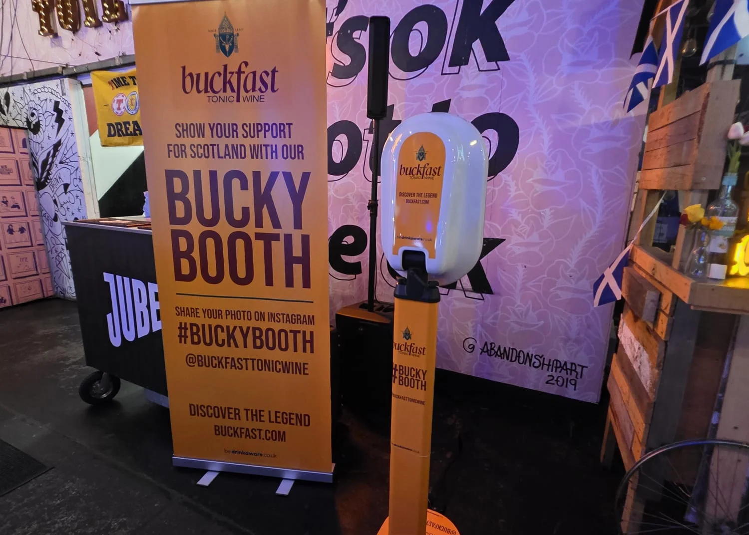 The Bucky Booth A.I. photo booth activation at Dockyard Social in Glasgow, showing the branded vinyl-wrapped booth unit alongside a custom orange pop-up banner displaying #BuckyBooth and @buckfasttonicwine branding.