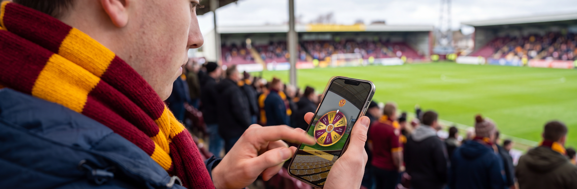 A football fan in a claret and amber scarf stands in the stand at a match, flicking his phone screen to spin a Motherwell FC Spin to Win prize wheel, with the pitch and crowd behind him.