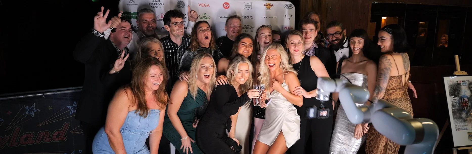 A large, energetic group of guests in formal attire laugh and pose together on a branded step-and-repeat backdrop at the Scottish Music Awards, with a robotic GlamCam arm visible in the foreground.