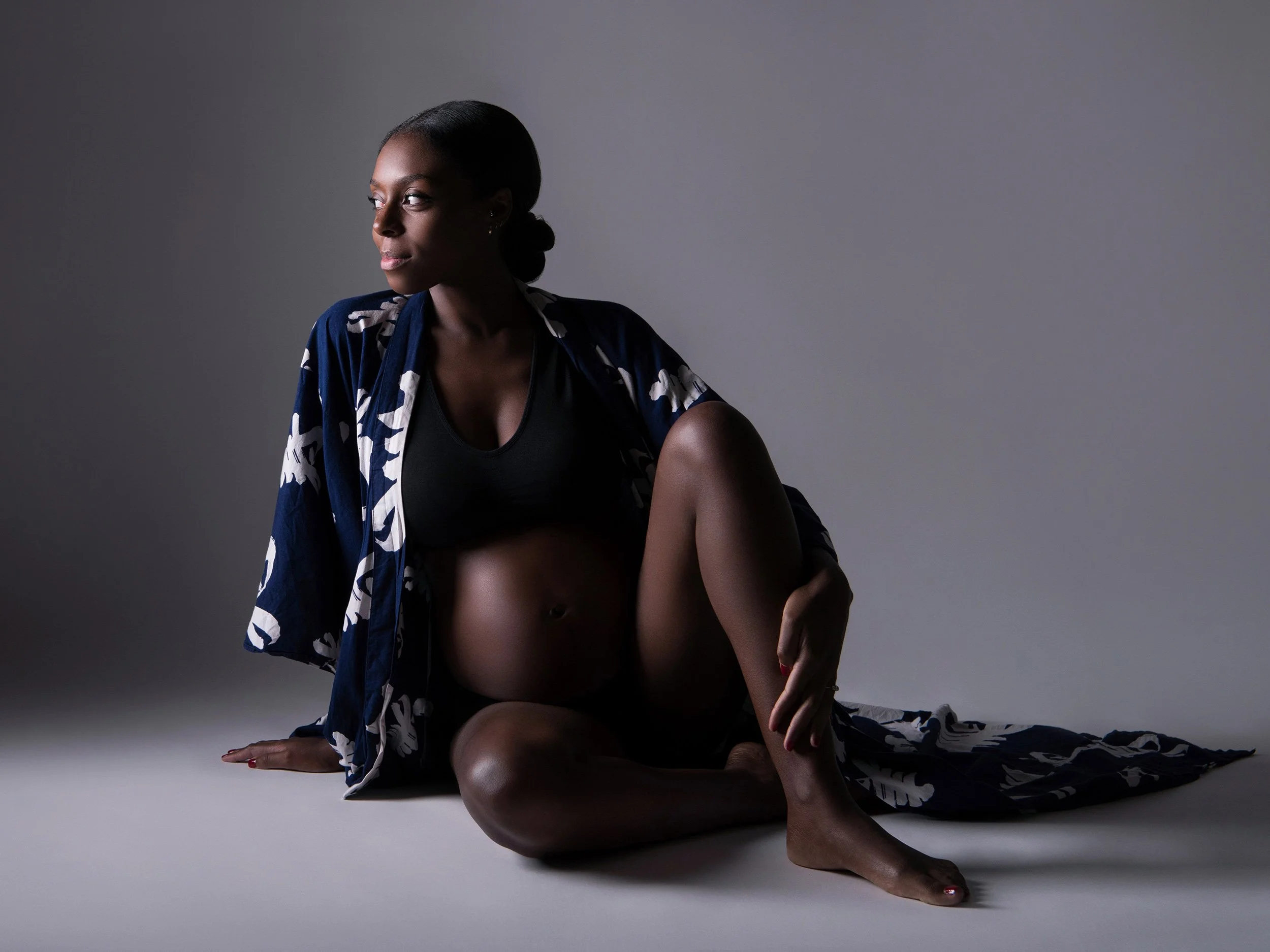 A woman sitting on the floor against a plain gray background, wearing a dark blue and white patterned robe, with her hair pulled back, looking to the side with a contemplative expression.