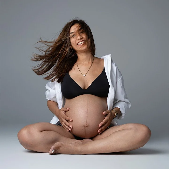 Pregnant woman sitting cross-legged in a black bra and open white shirt, smiling during a maternity photoshoot in a London studio.