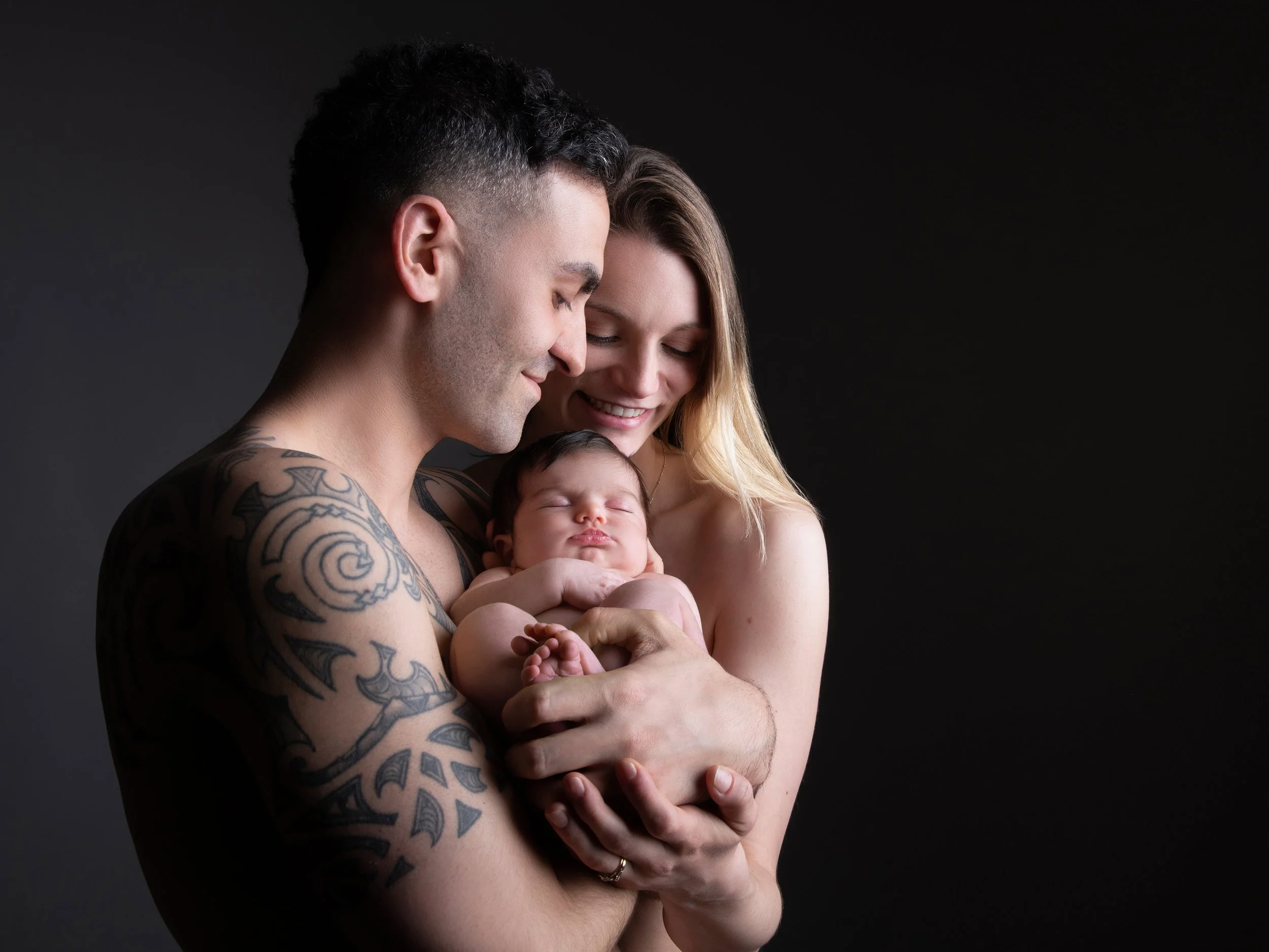 A happy couple holding a newborn baby against a dark background.