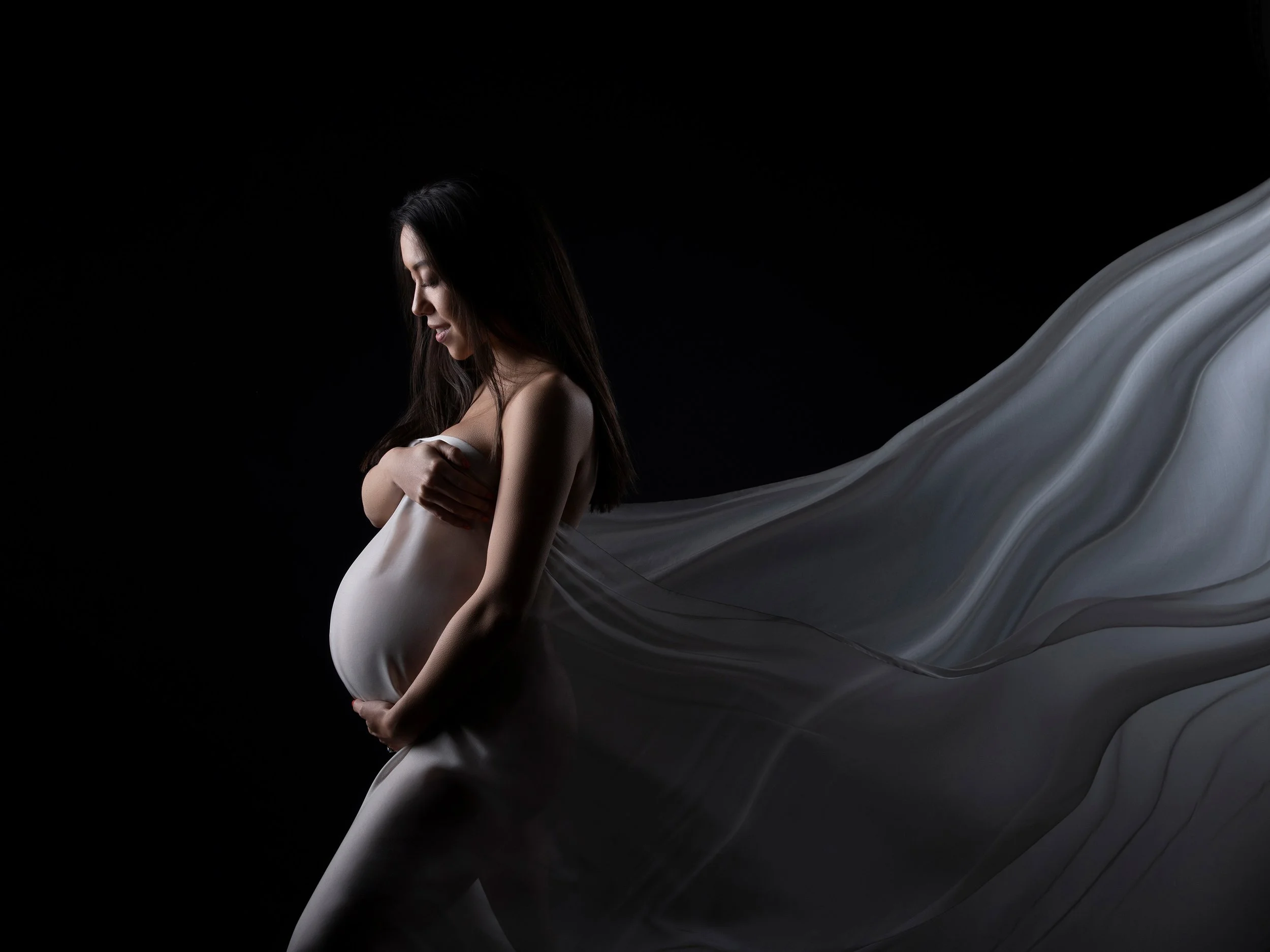 Fine-art maternity portrait of a pregnant woman with long dark hair, cradling her belly with her eyes closed as white fabric flows around her against a dark studio background.