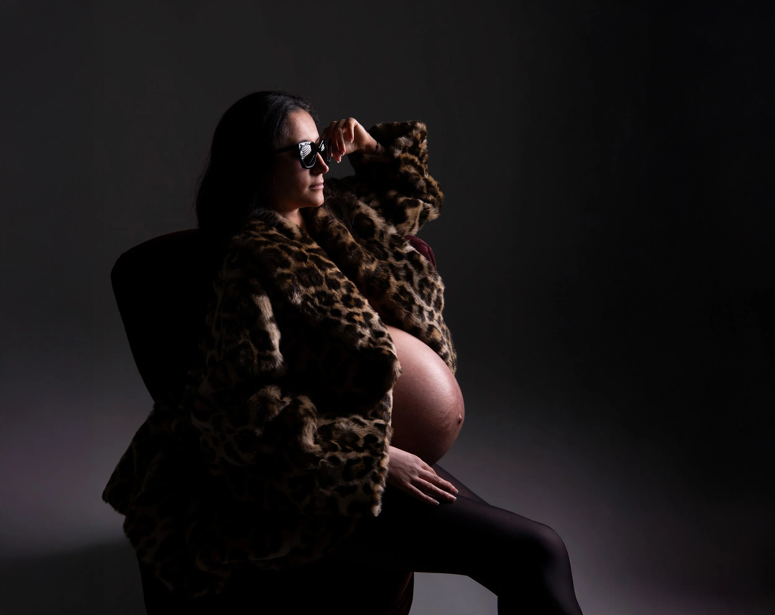 A woman wearing a leopard-print coat and sunglasses, with long dark hair, sitting on a dark surface, in a dark studio setting, with her hand on her pregnant belly.