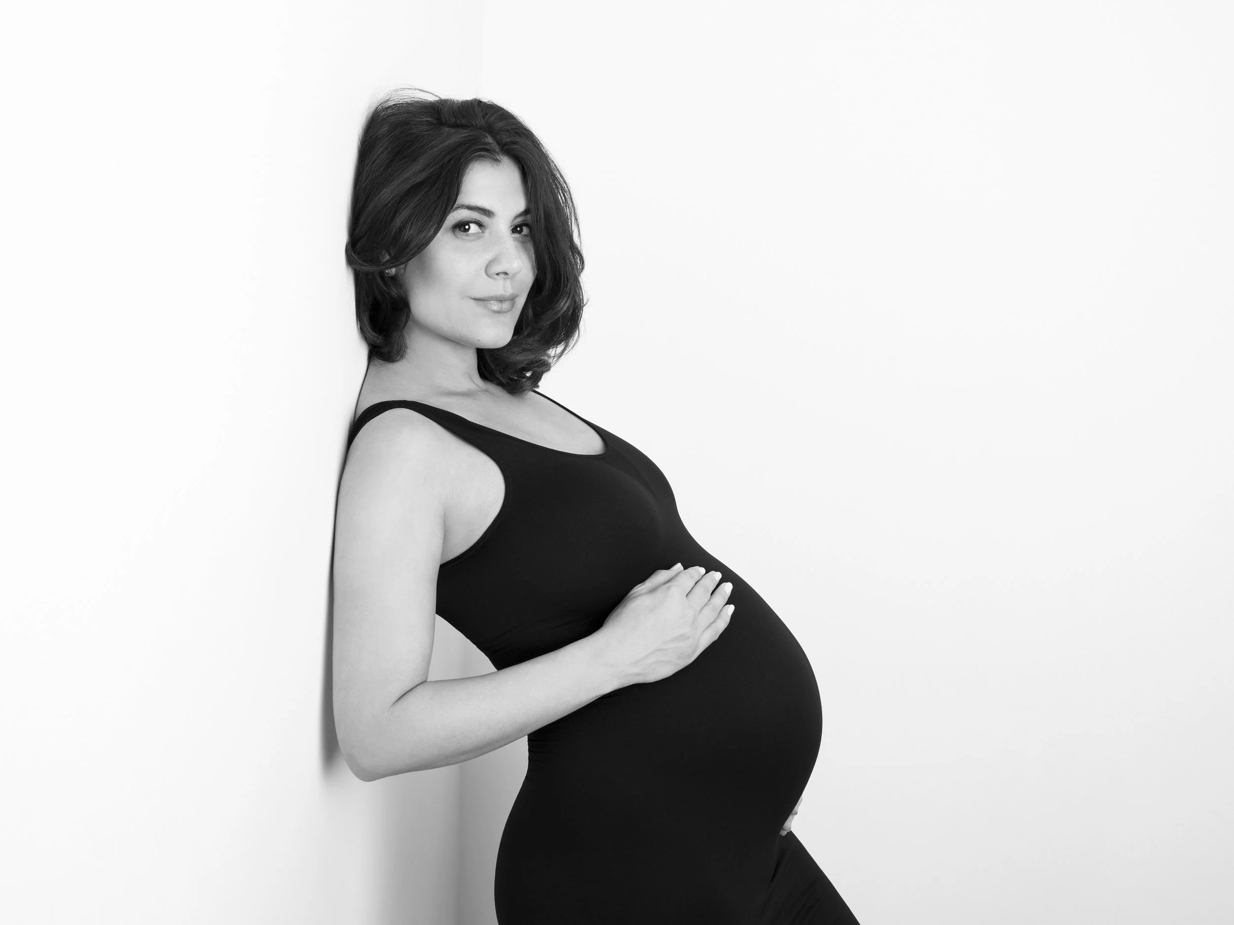 Black and white photo of a pregnant woman with shoulder-length hair, wearing a sleeveless top, standing against a plain wall, gently holding her belly, with a serene expression.