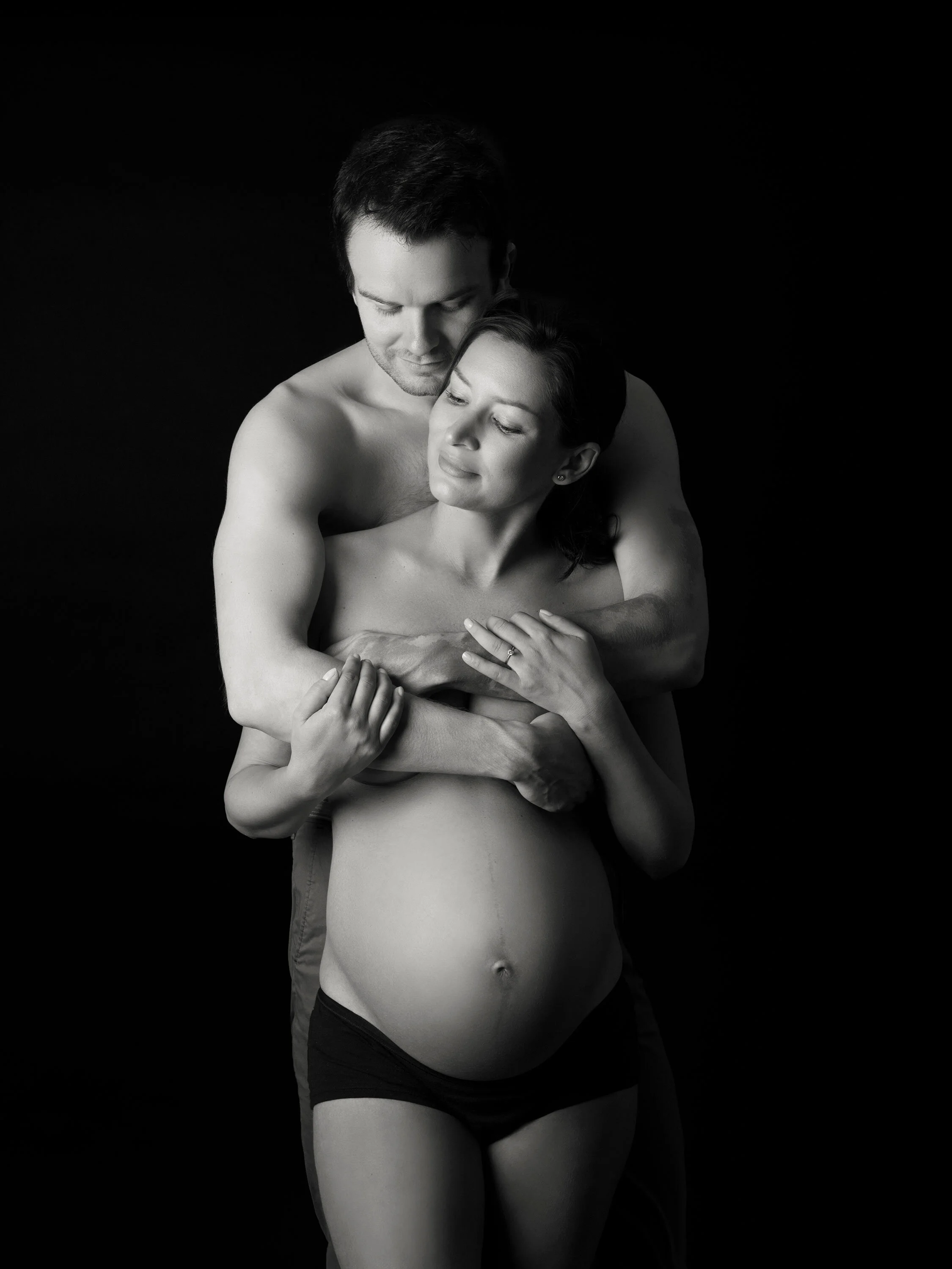 Black and white photo of a man and woman embracing. The woman is wearing underwear, and the man is shirtless. They are holding each other lovingly against a dark background.