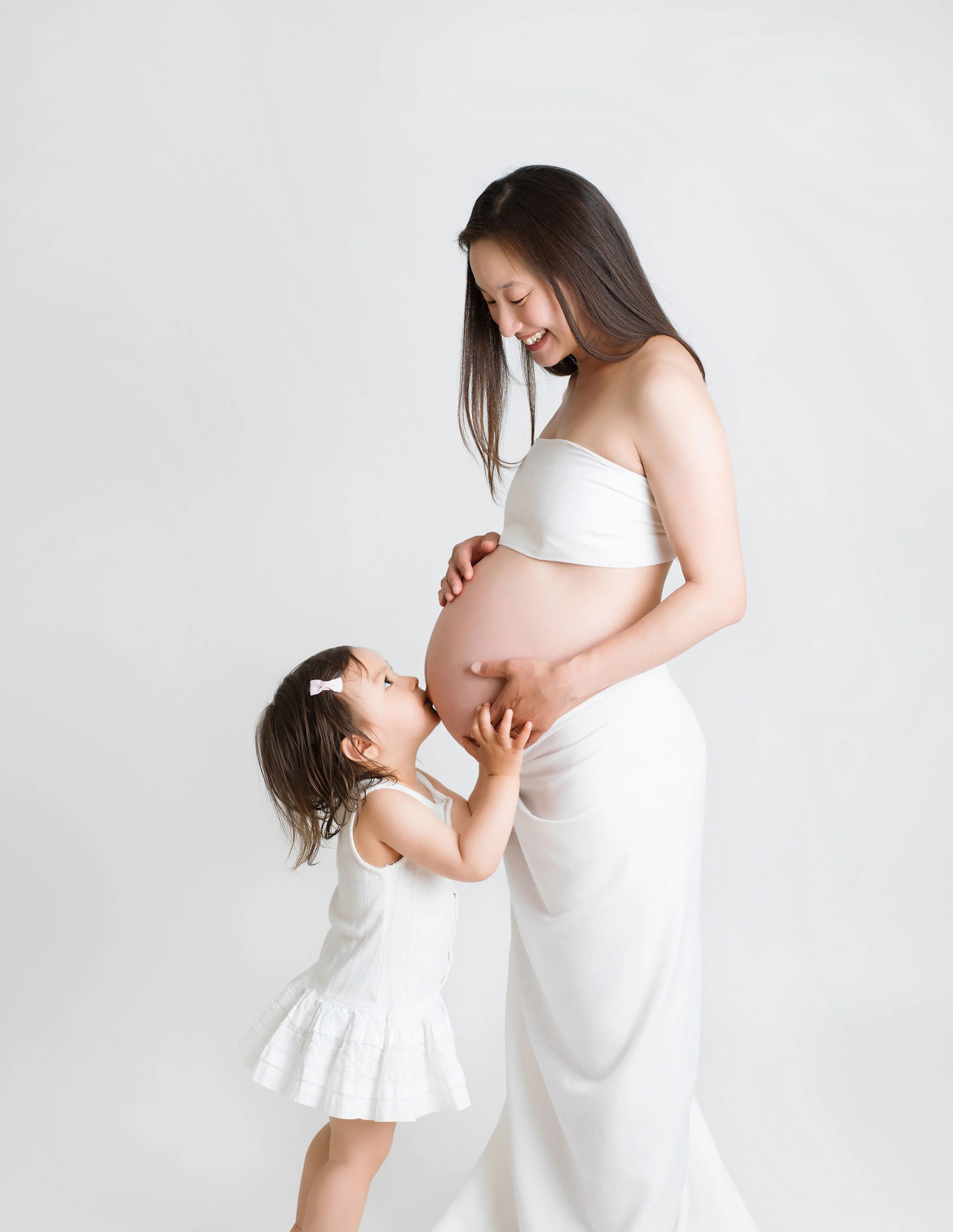 Maternity portrait of a pregnant woman dressed in white, smiling down at her young daughter as she touches her belly in a bright, minimalist studio.