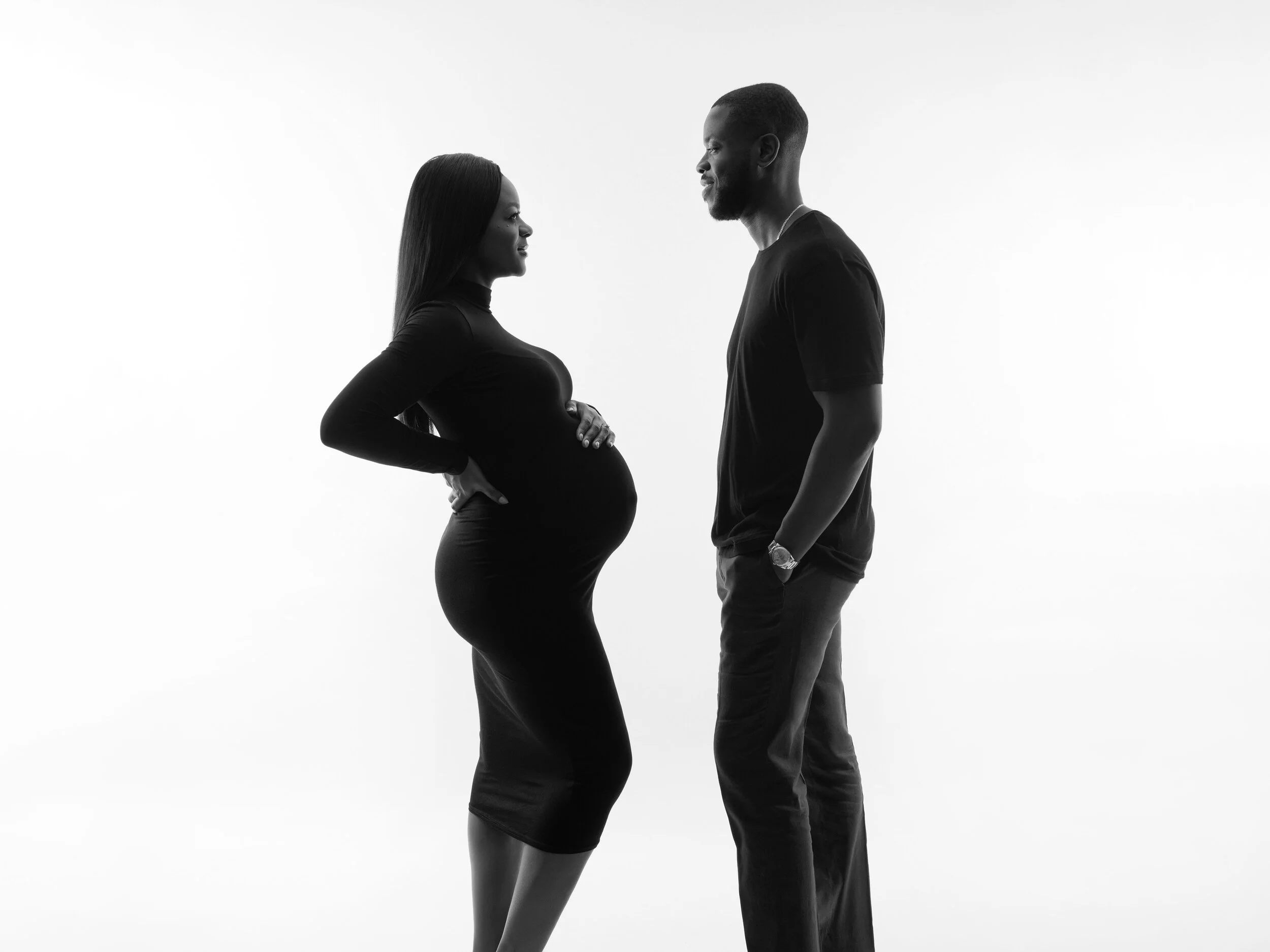 A silhouette portrait of a pregnant woman and a man facing each other in a black-and-white photo, standing close with the woman holding her belly and the man with his hands in his pockets.