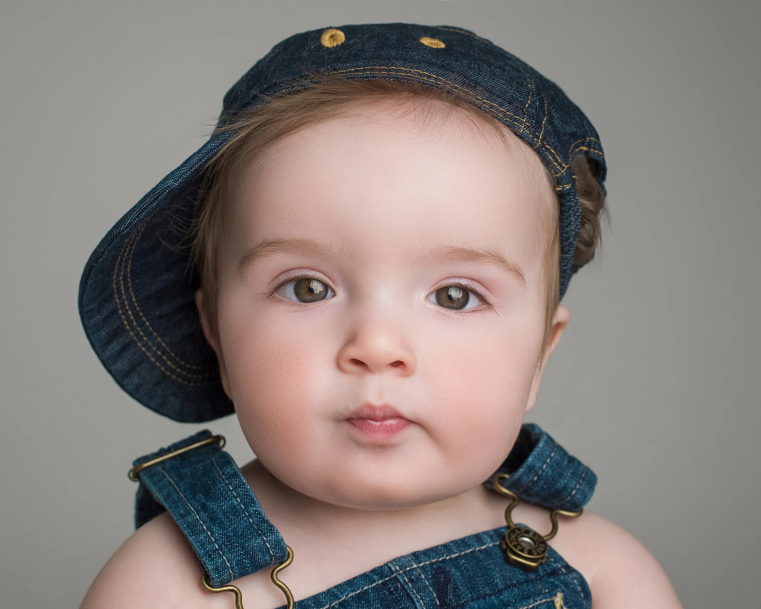 Close-up of a young boy with light brown hair and brown eyes, wearing a denim hat and matching denim suspenders, against a plain gray background.
