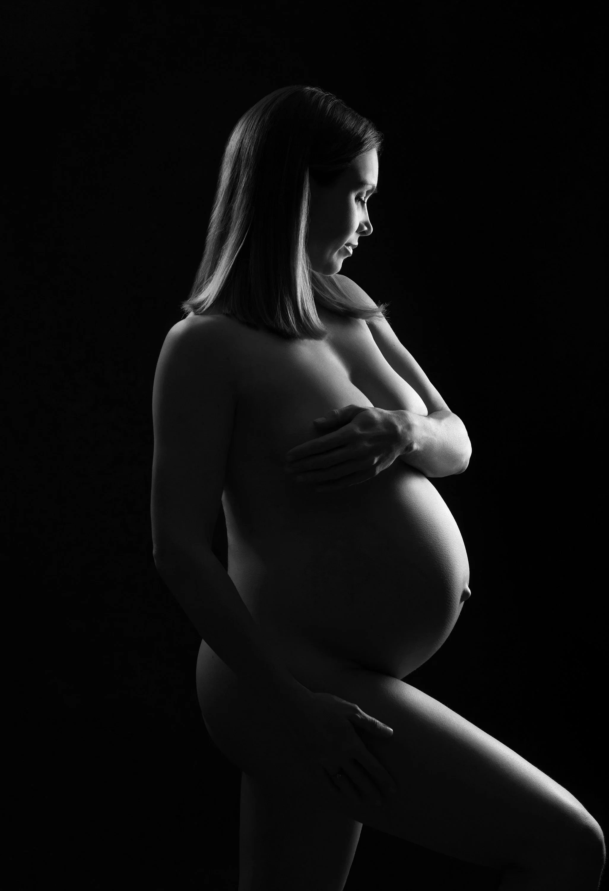 Black-and-white maternity portrait of a pregnant woman cradling her belly, with soft lighting highlighting her profile against a dark background.