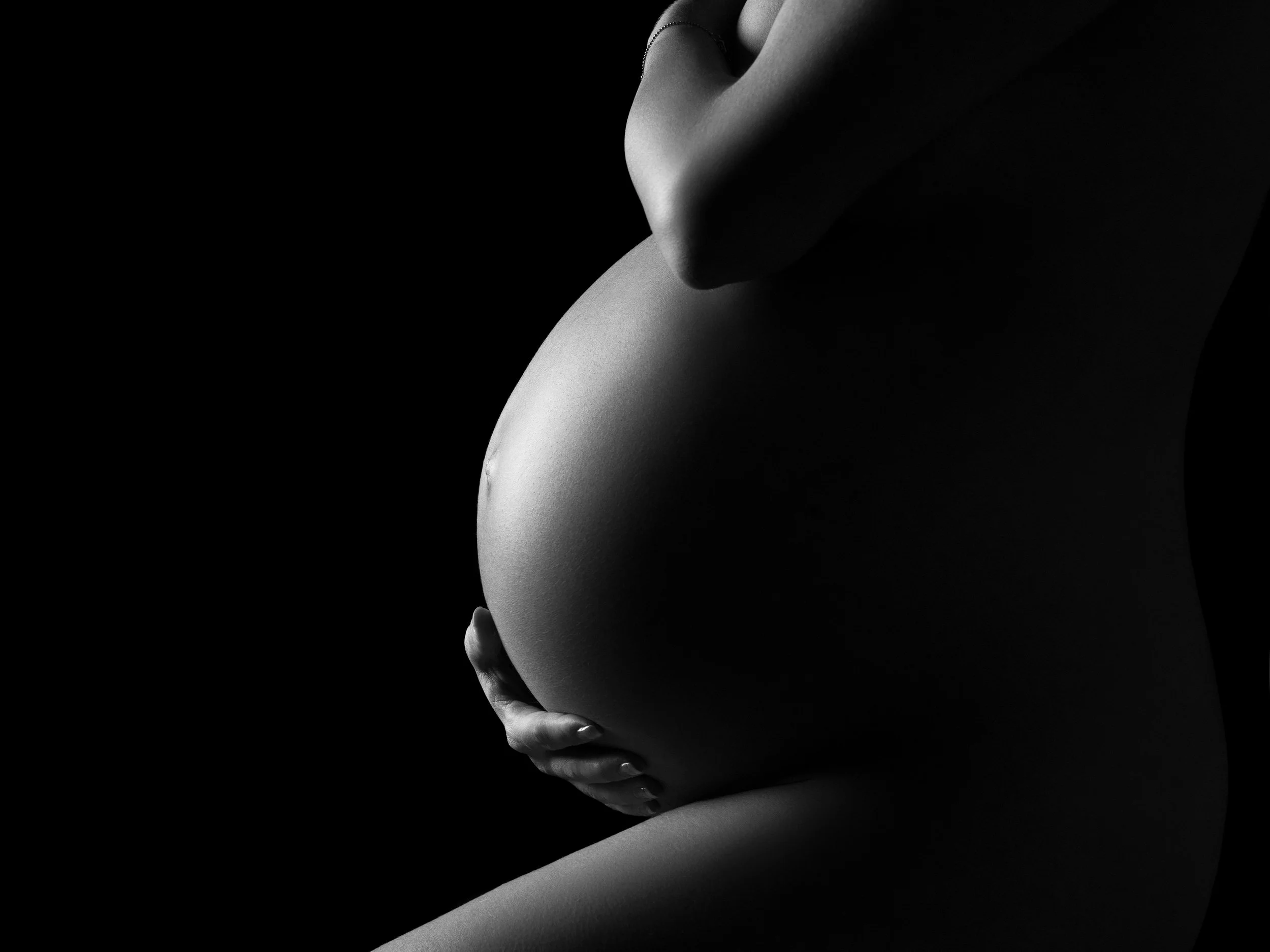 Naked pregnancy photography in a West London photography studio. A black and white close-up of a woman's body, with one hand cradling her pregnant belly.