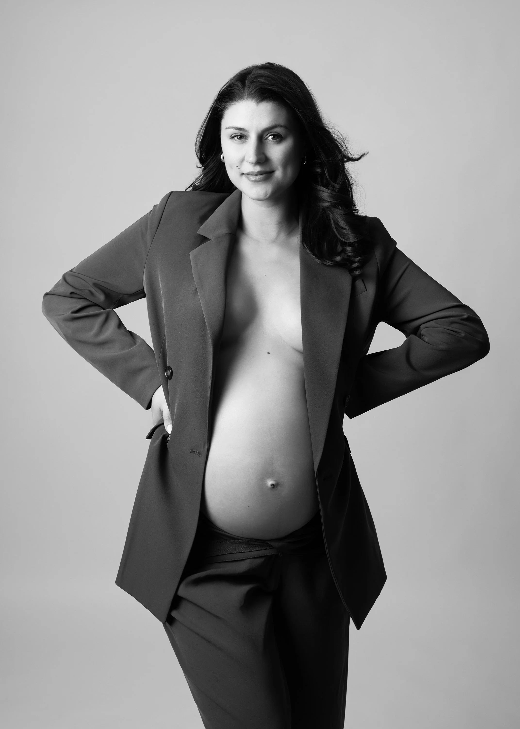 Black and white maternity portrait of a confident mum-to-be in a tailored blazer, hands on hips, in a London studio