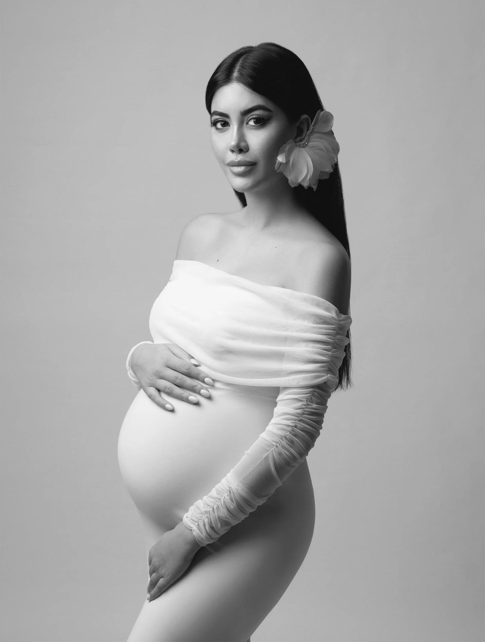 Black-and-white editorial maternity portrait of Rojin Tufan wearing a long-sleeved white dress and statement flower earring against a light studio background.
