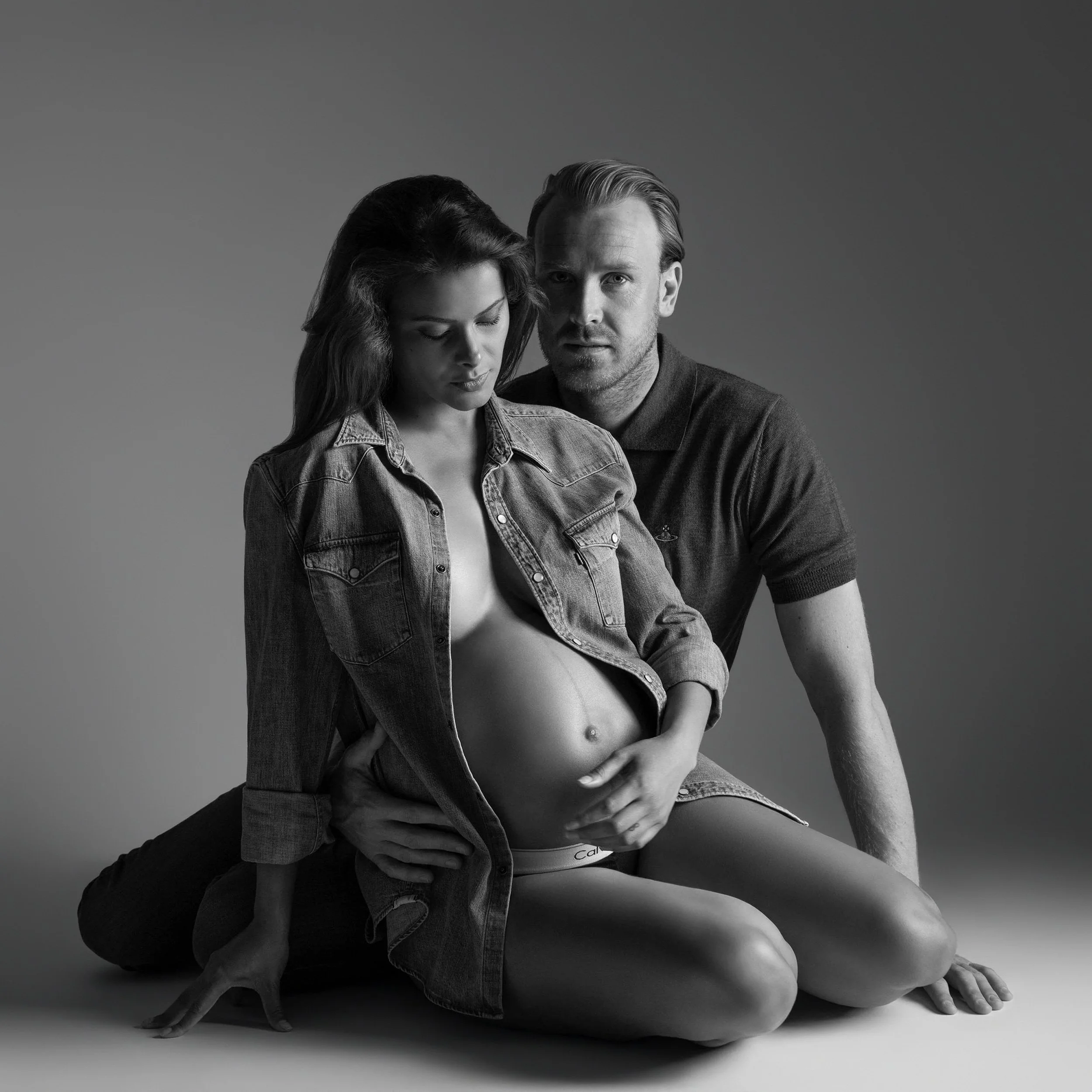 Black-and-white couples maternity portrait of a pregnant woman in a denim shirt seated on the floor, with her partner behind her and his hands resting on her waist, against a plain grey studio background.