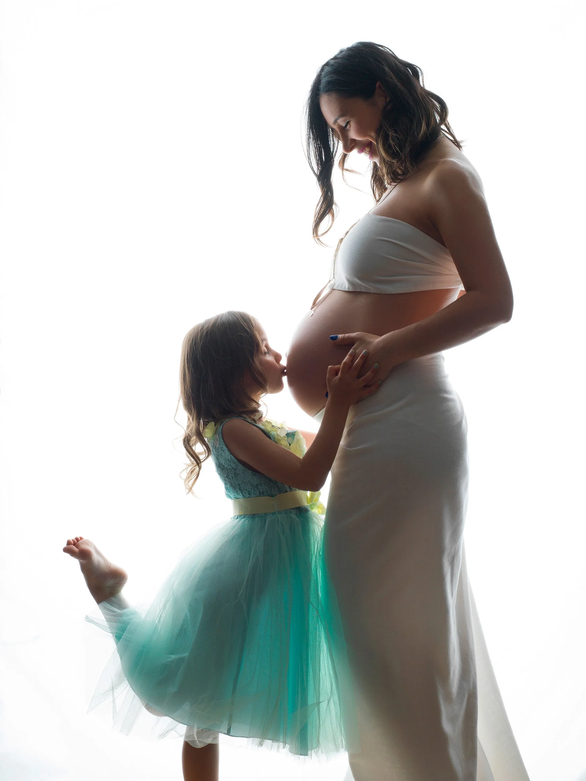 A pregnant woman with dark hair in a white strapless dress smiling at a young girl with long hair in a colorful dress and tulle skirt, both embracing her belly against a bright white background.