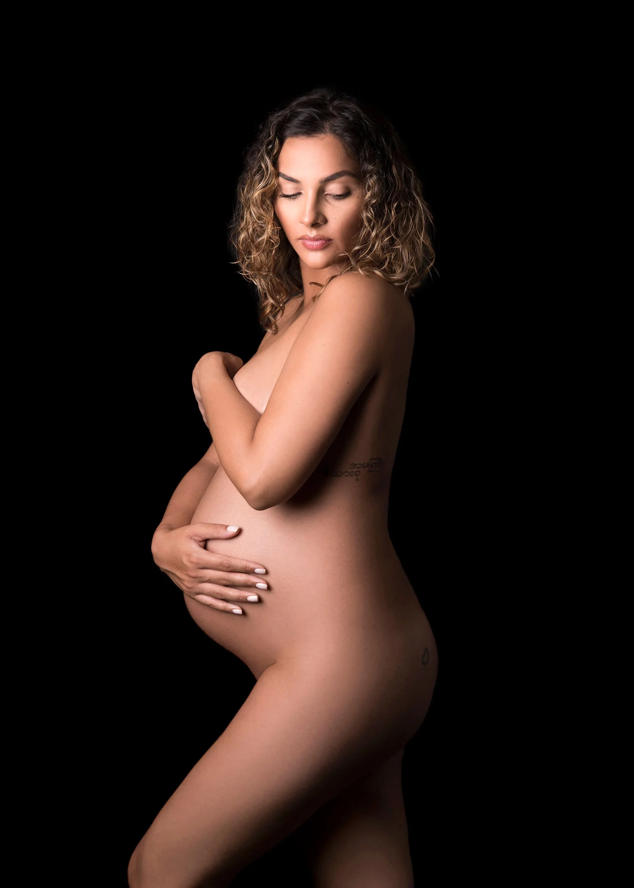 Nude pregnant woman with curly hair and tattoos posing against a black background in an artistic maternity portrait.
