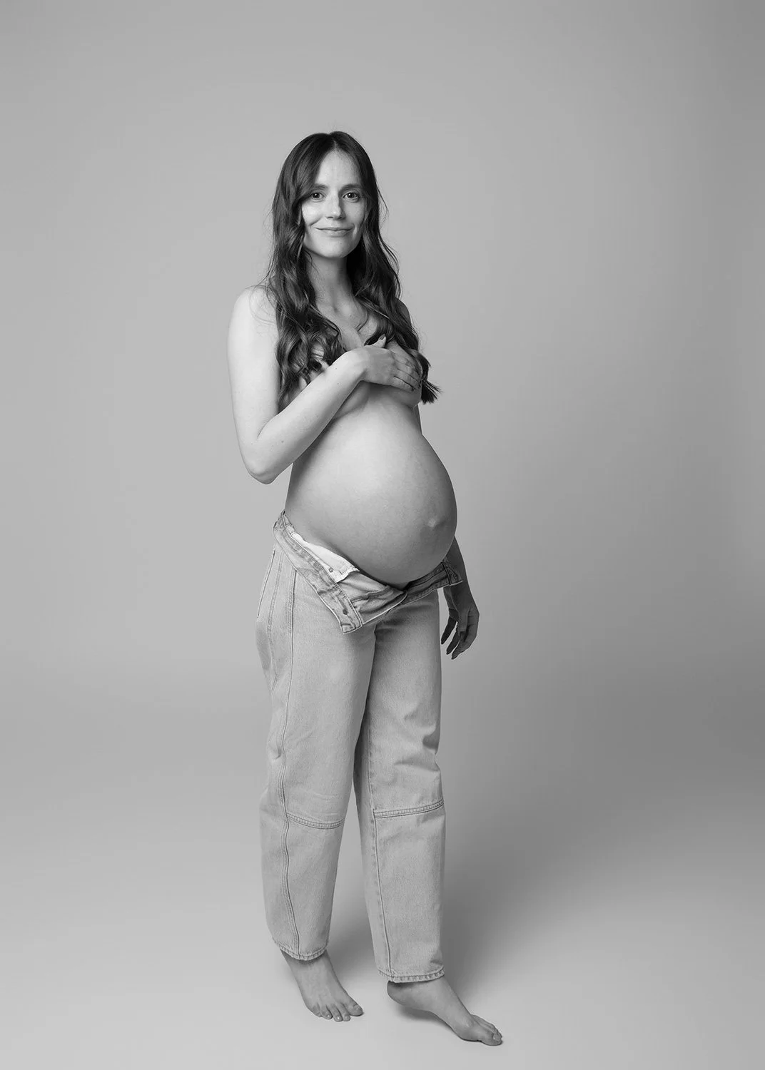 Black and white photo of a pregnant woman with long hair, wearing jeans, cover her chest with her hand, standing on a plain background.