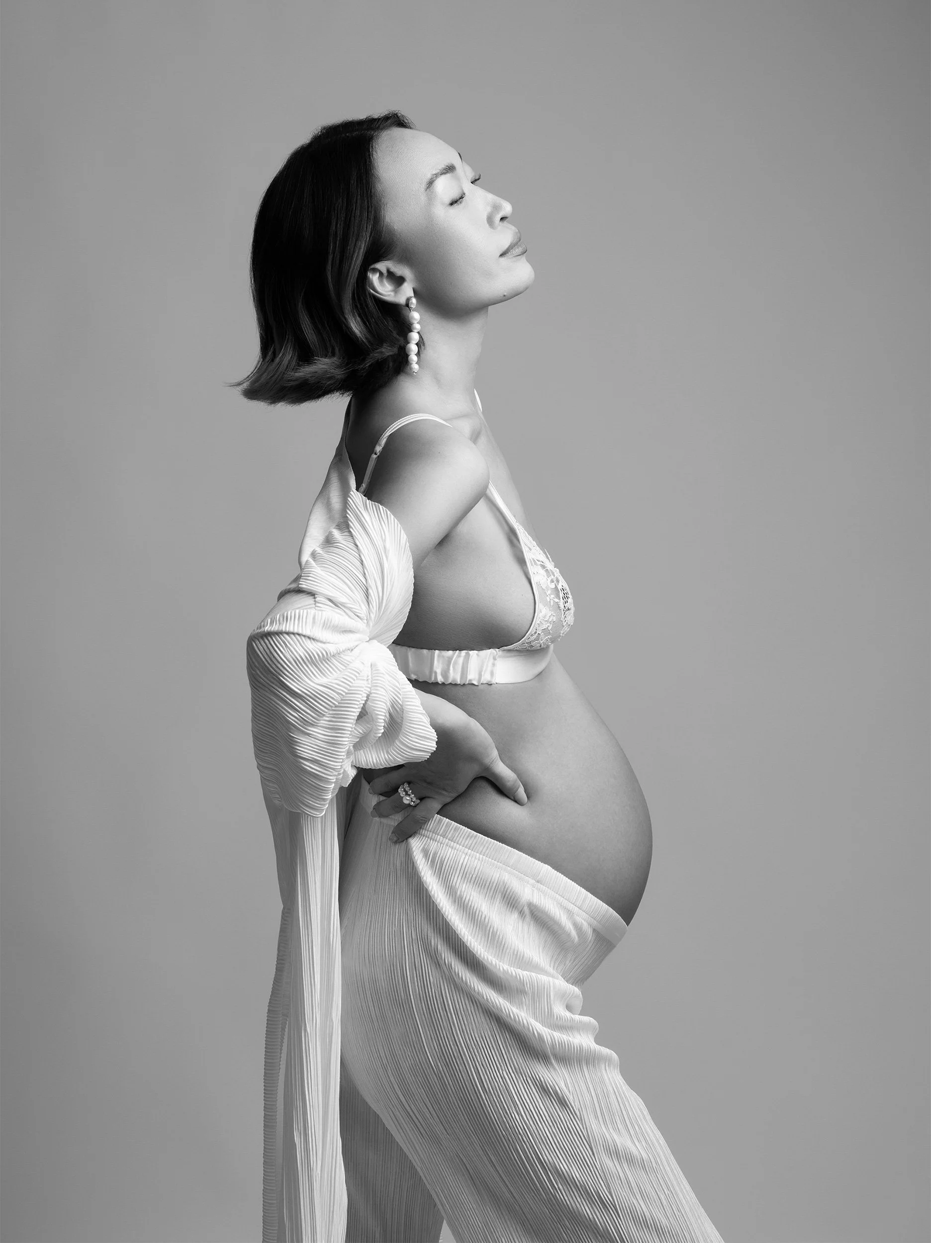 Black and white photograph of a pregnant woman with shoulder-length hair, wearing earrings, a lace bra, and flowing fabric, with her eyes closed and head tilted back, posing against a plain background.