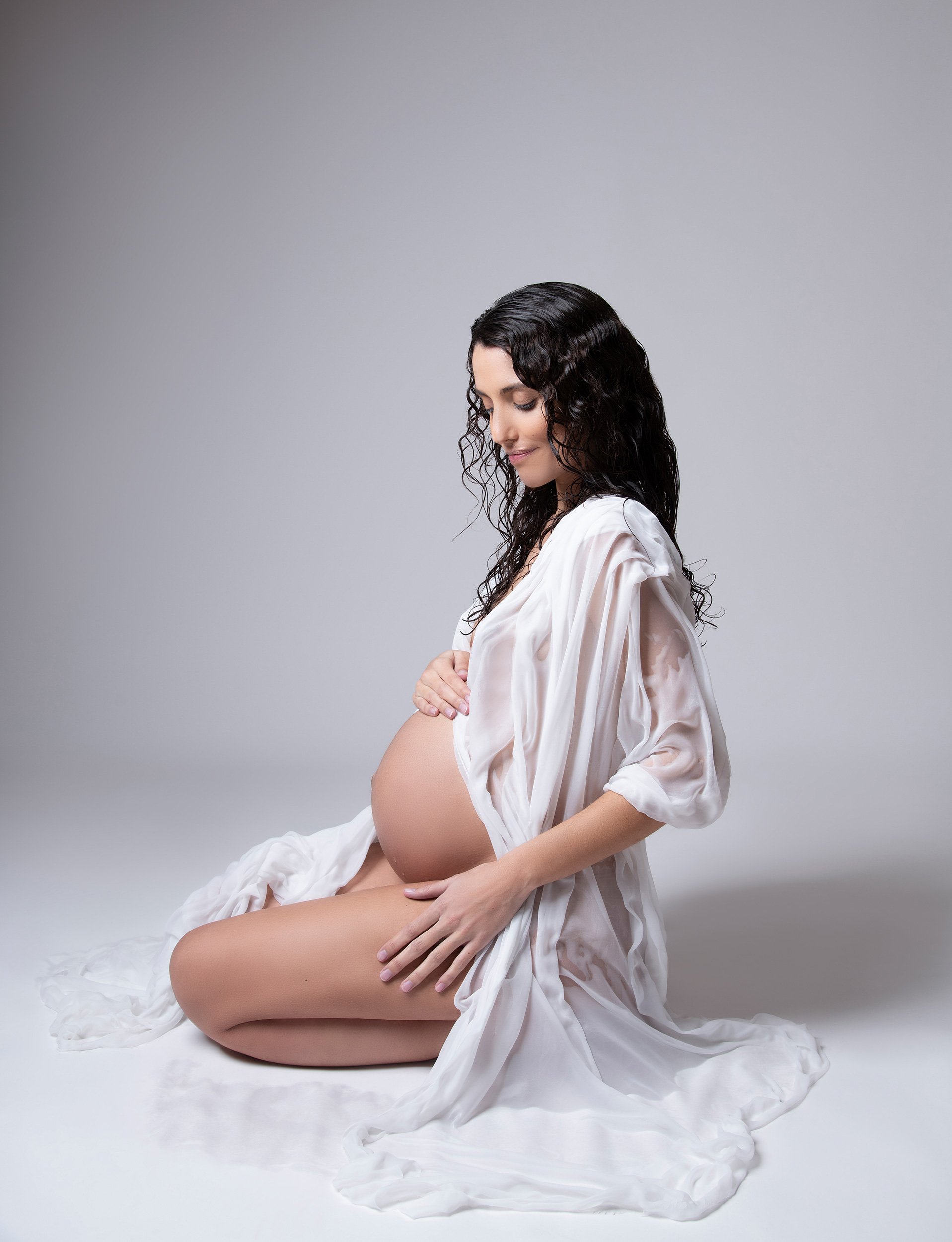A pregnant woman with long curly black hair, wearing a white satin robe, sitting on the floor, and cradling her belly with both hands, against a plain gray background.