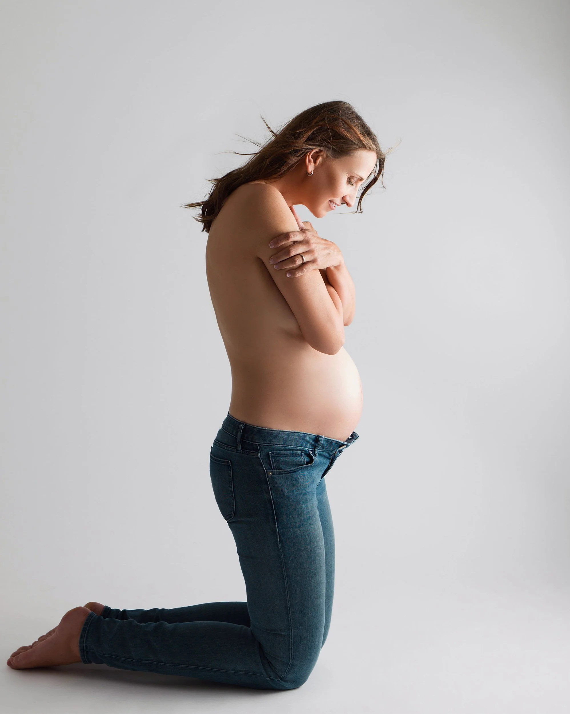 A pregnant woman with brown hair in a yoga pose, kneeling on the floor with arms crossed over her chest and looking downward, against a plain gray background.