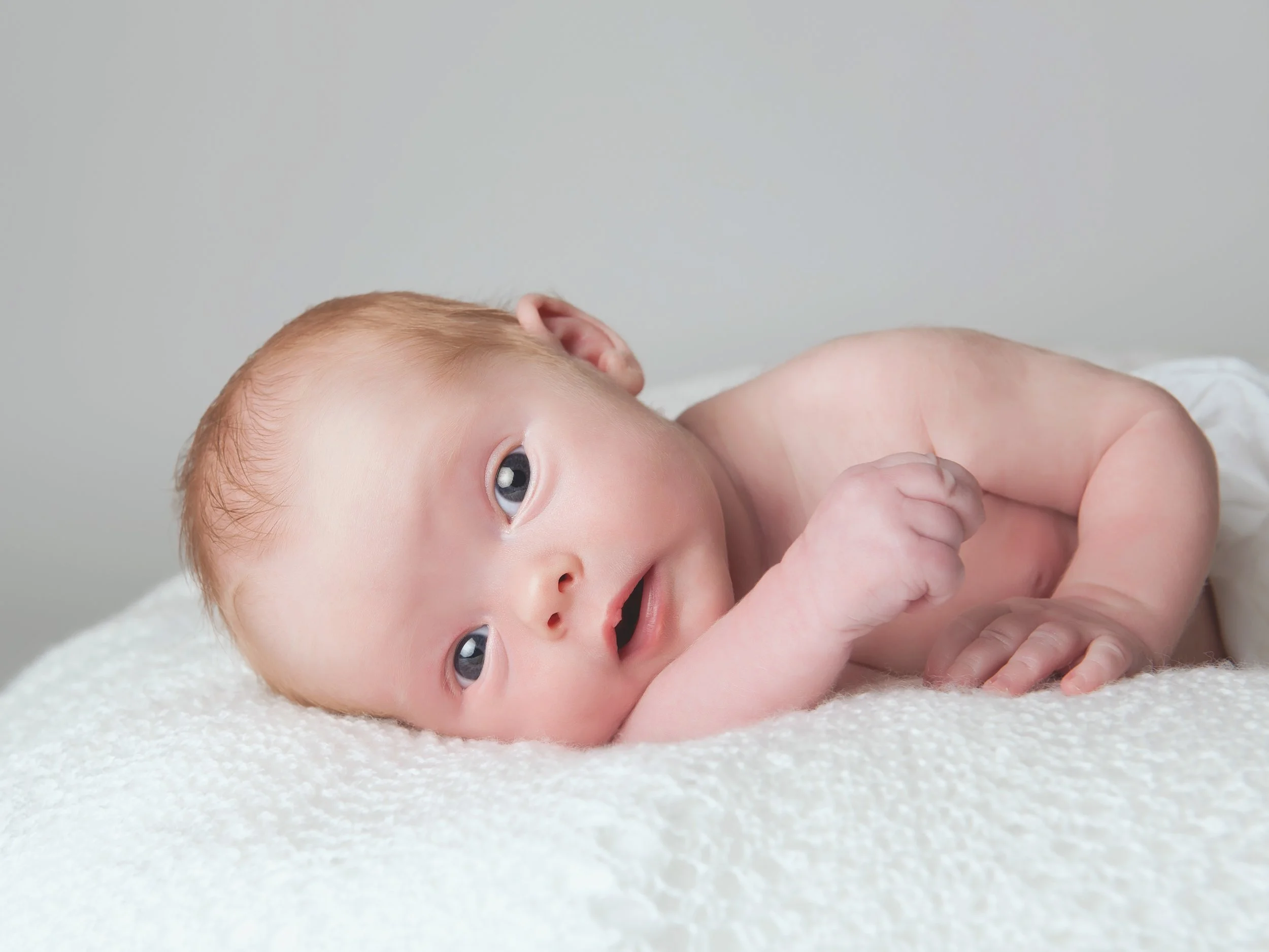 Close-up portrait by a London newborn photographer of a blue-eyed baby lying on soft white fabric and looking towards the camera.