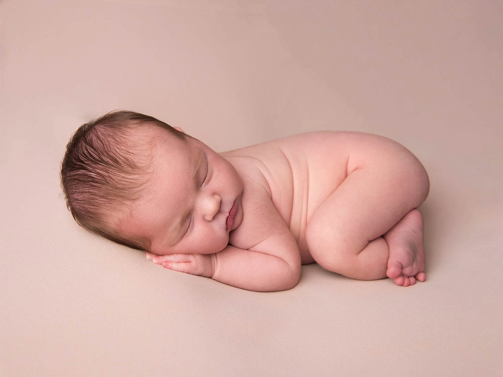 A baby with dark hair sleeping on a beige surface, curled up with hands under the cheek, facing to the side.