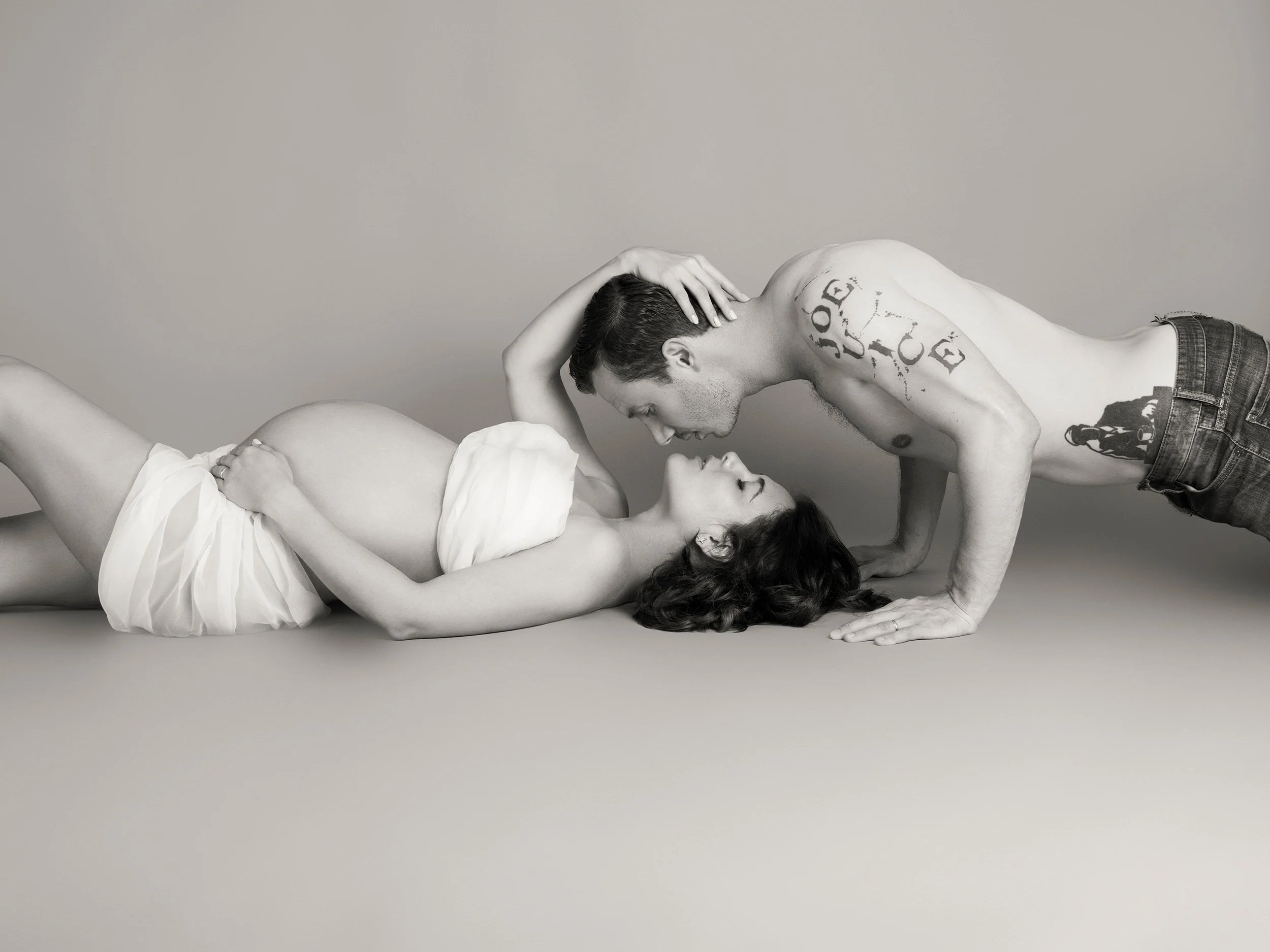 A black-and-white photo of a shirtless man leaning over a woman lying on her back. The man is wearing jeans and has a tattoo on his shoulder. The woman is draped in a white cloth and has long dark hair. They are on a plain surface against a neutral b