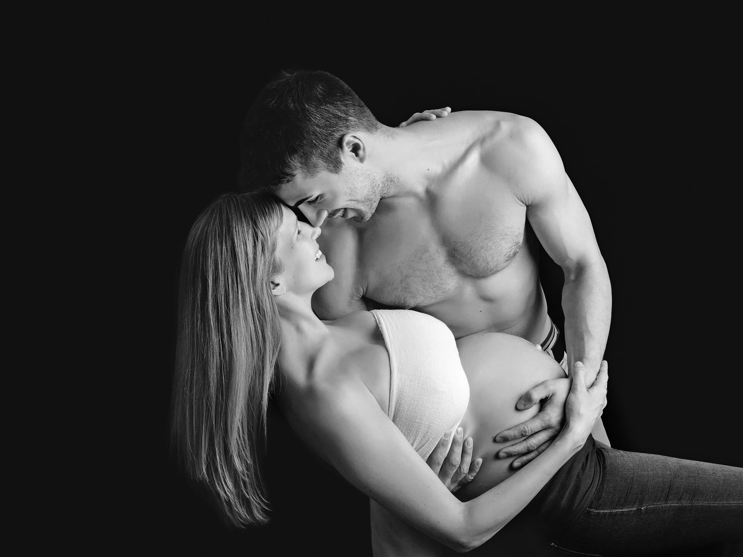 A black and white photo of a man and woman in an intimate pose, with their foreheads touching and smiling, against a dark background.