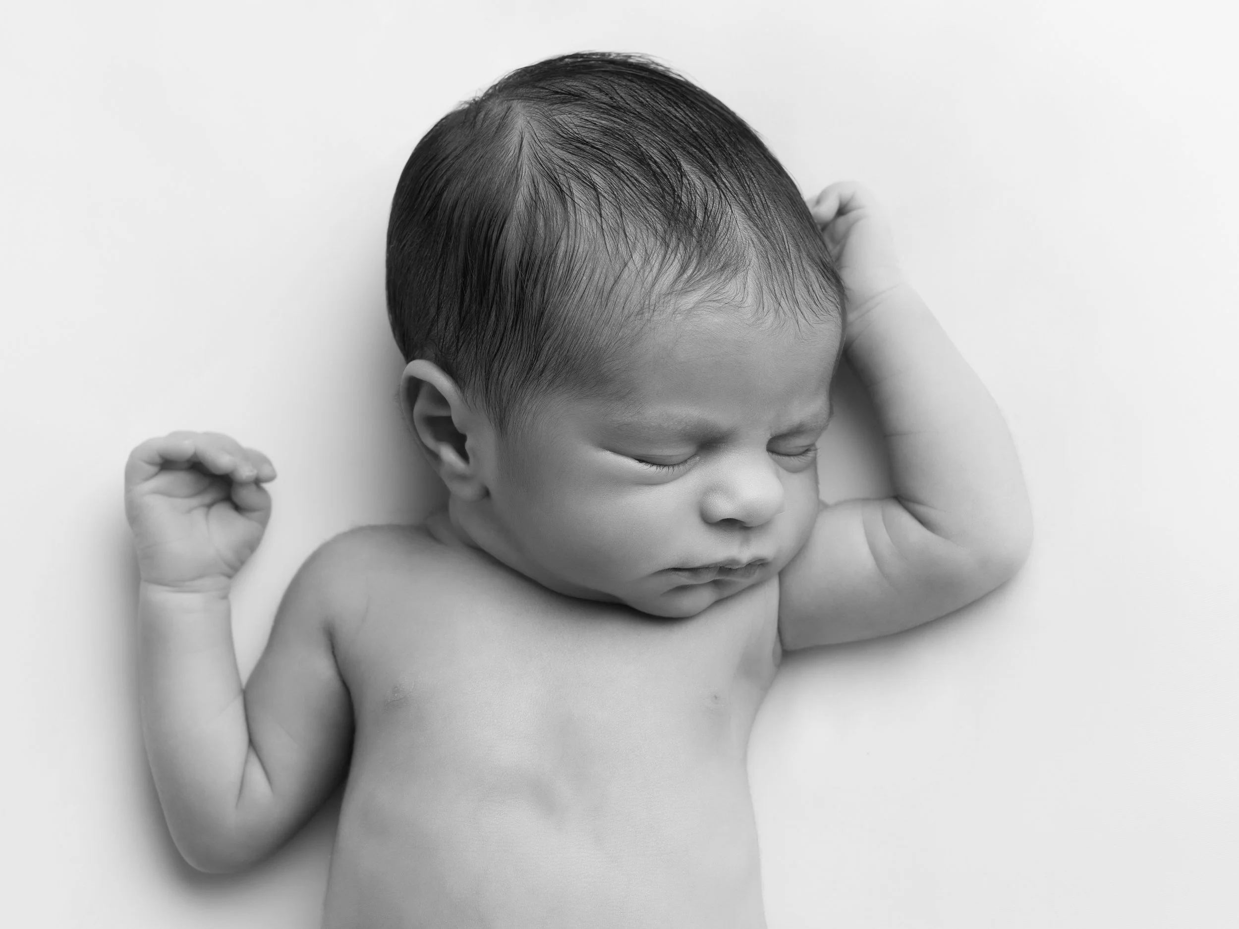 Black and white photo of a sleeping baby with closed eyes, outdoors, with one arm bent and near the head, and the other arm bent with a fist near the chest.