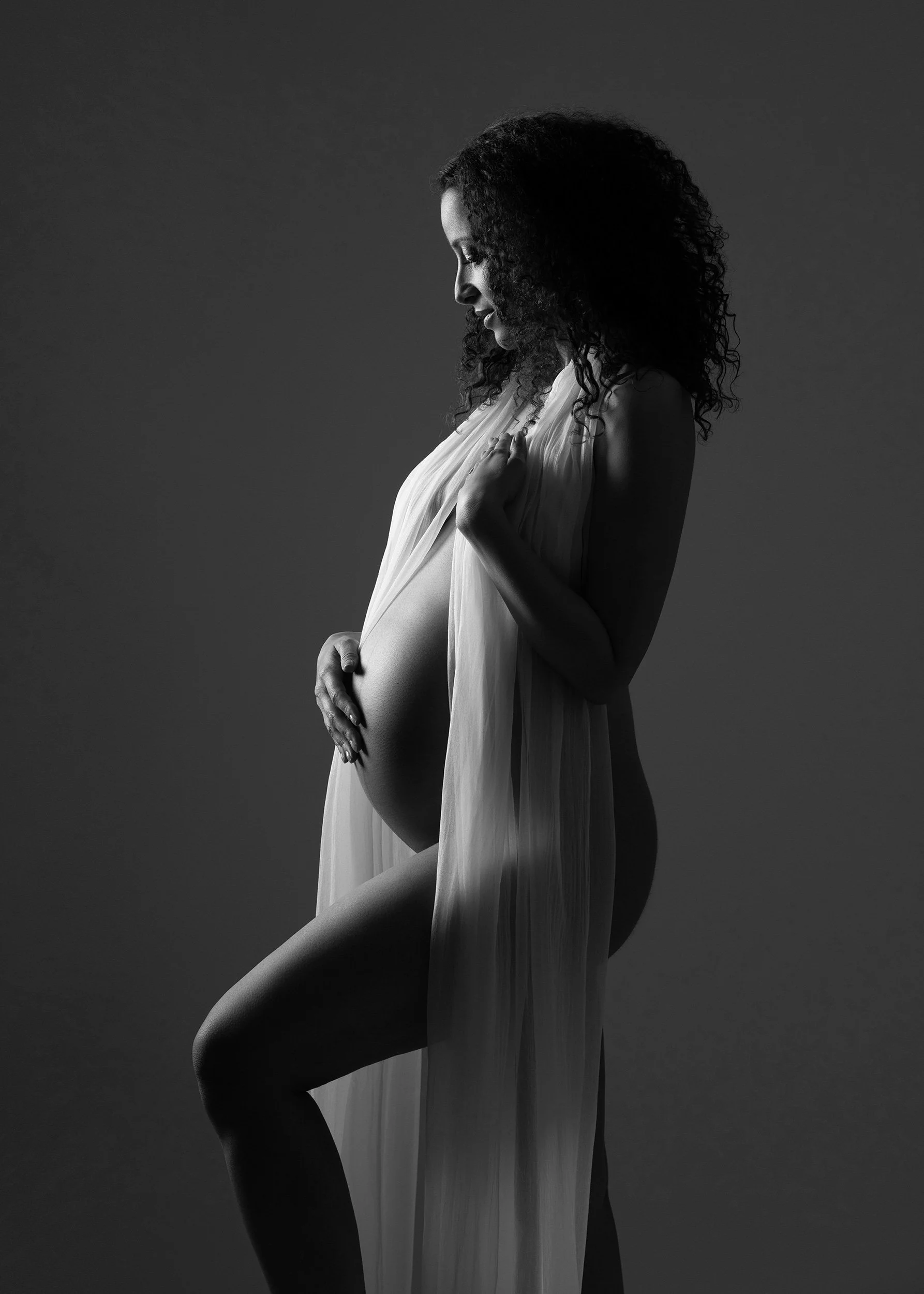 Black and white photograph of a pregnant woman draped in flowing fabric, gently holding her belly with both hands, standing in profile against a dark background.