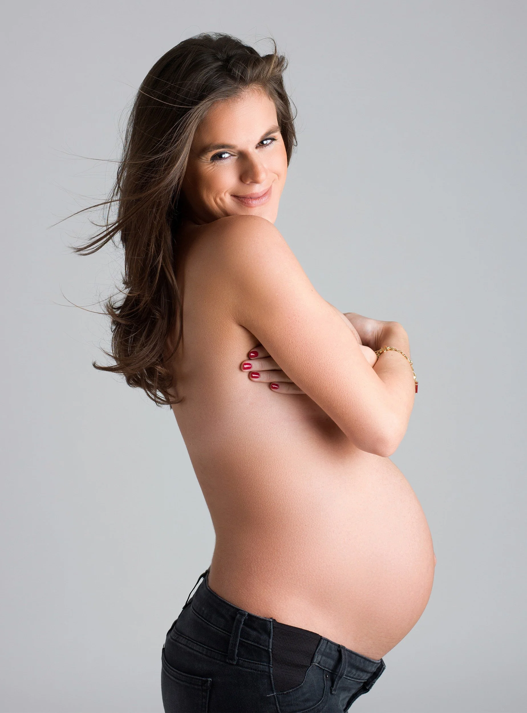 A pregnant woman with long brown hair smiling and crossing her arms over her chest, wearing dark jeans and a gold bracelet, against a plain gray background.