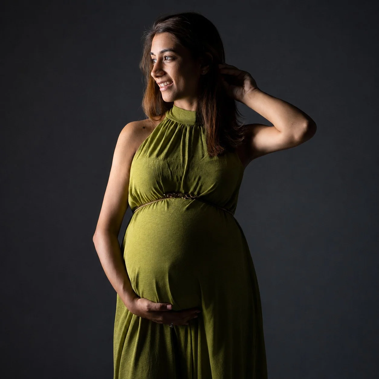 A smiling pregnancy photograph of a woman in a green dress, holding her belly with one hand and touching her hair with the other, standing against a dark background.