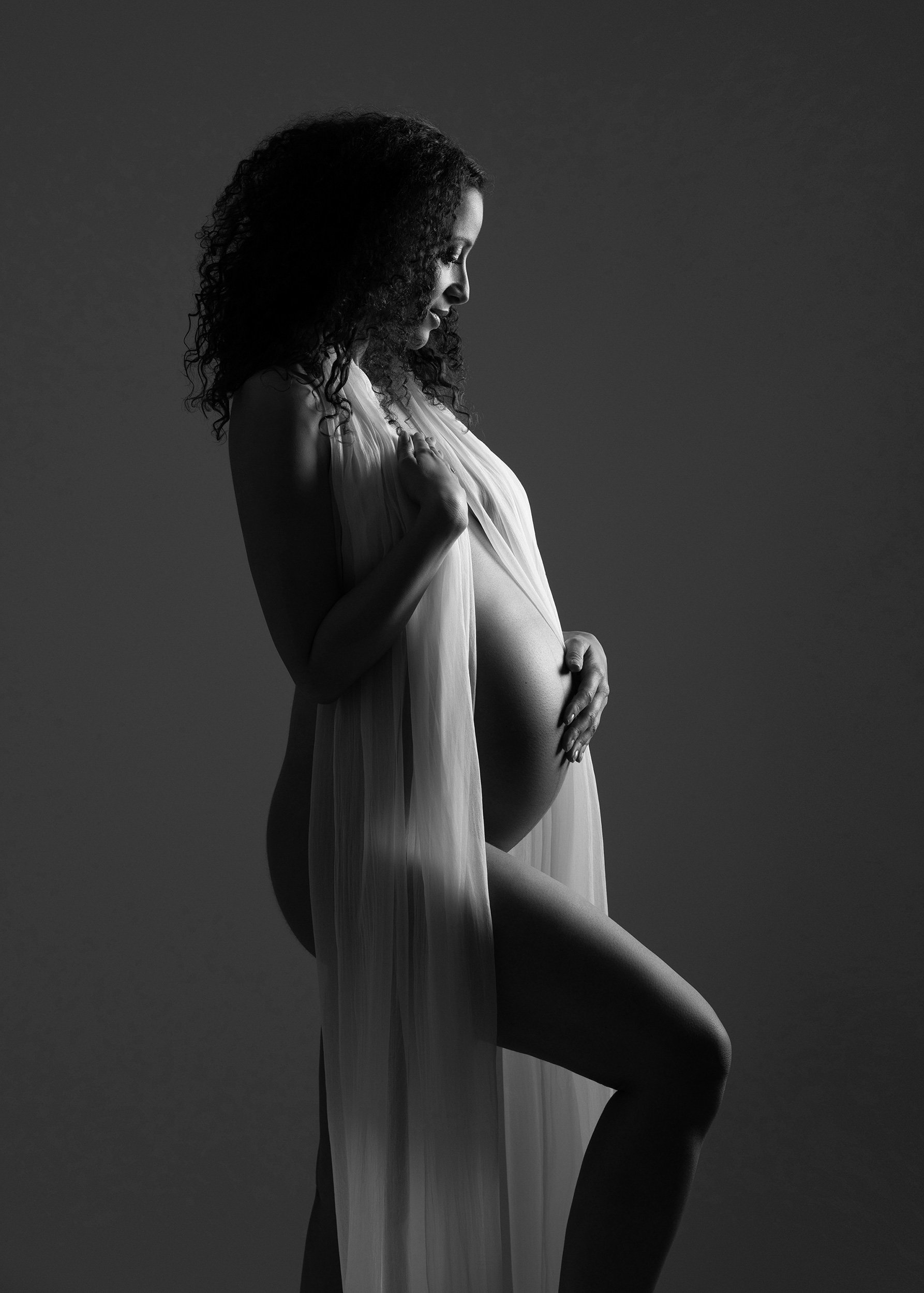 Black-and-white fine-art maternity portrait of a pregnant woman with curly hair, draped in sheer flowing fabric and holding her belly against a dark studio background.