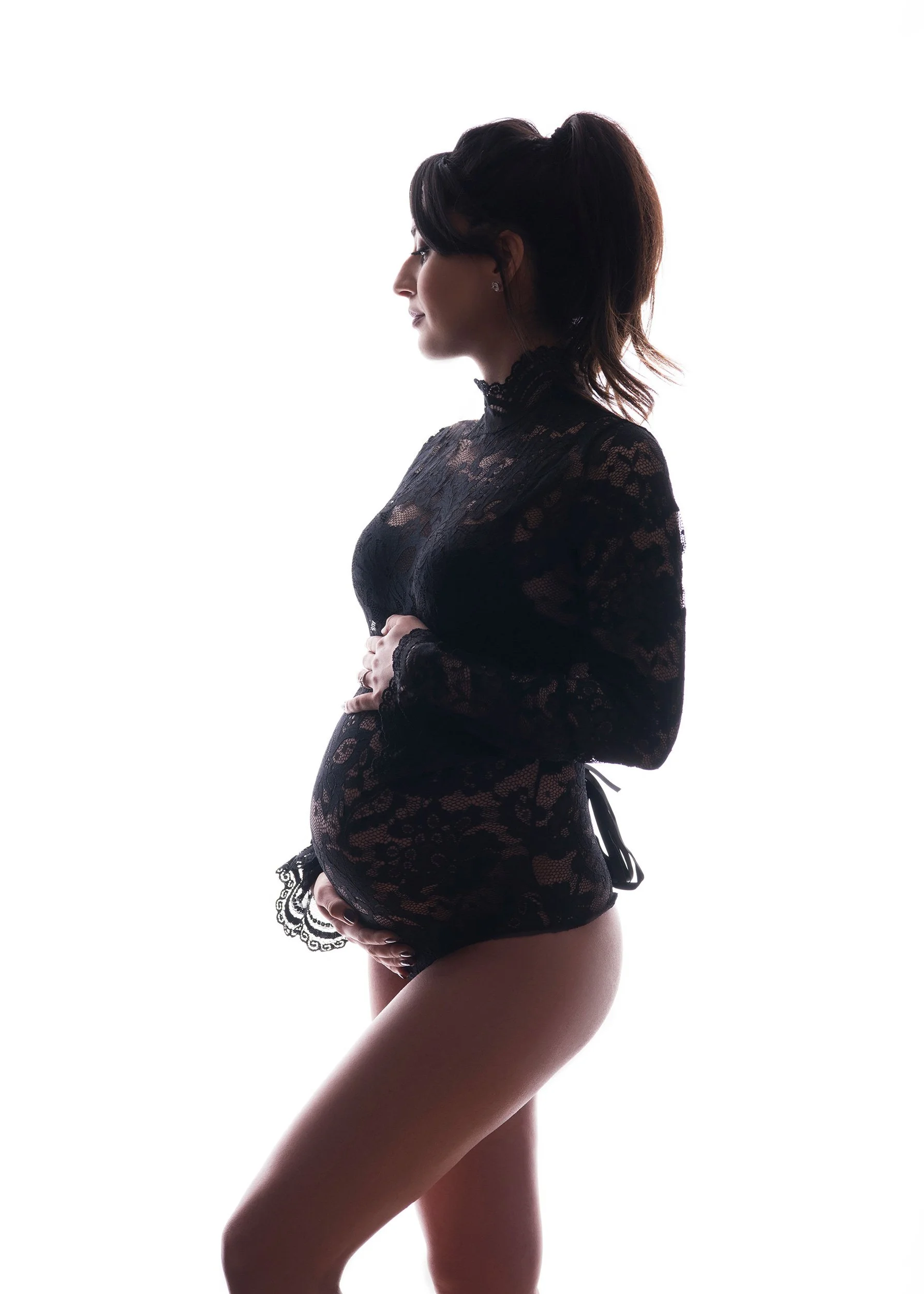 Profile of a woman in black lace lingerie with her eyes closed and hands on her abdomen, standing in front of a white background.