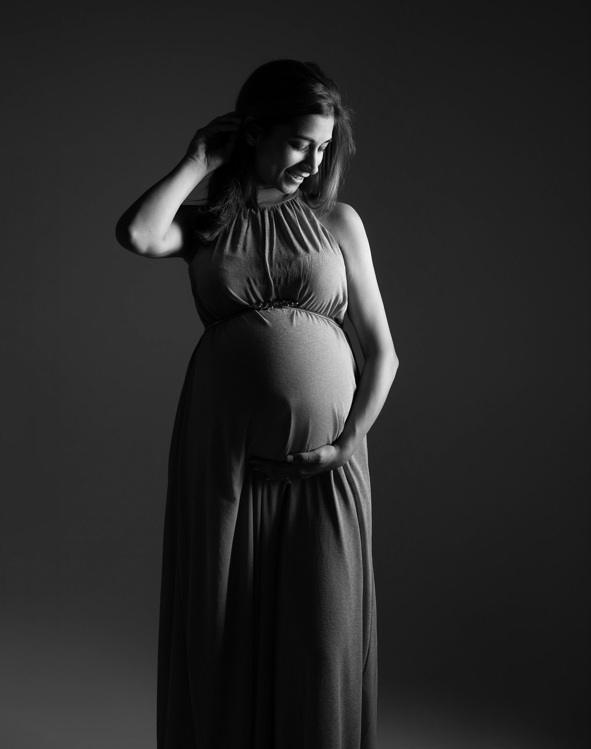 Black and white maternity portrait of a pregnant woman in a sleeveless dress, smiling and gently holding her bump against a dark studio background.