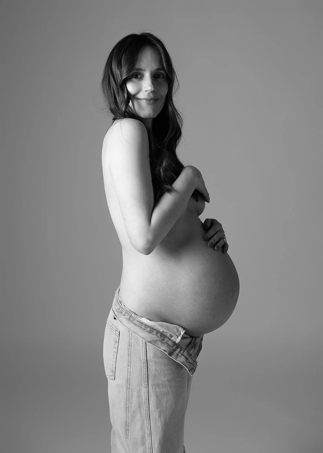 Black and white pregnancy semi-nude studio portrait of a woman wearing jeans.